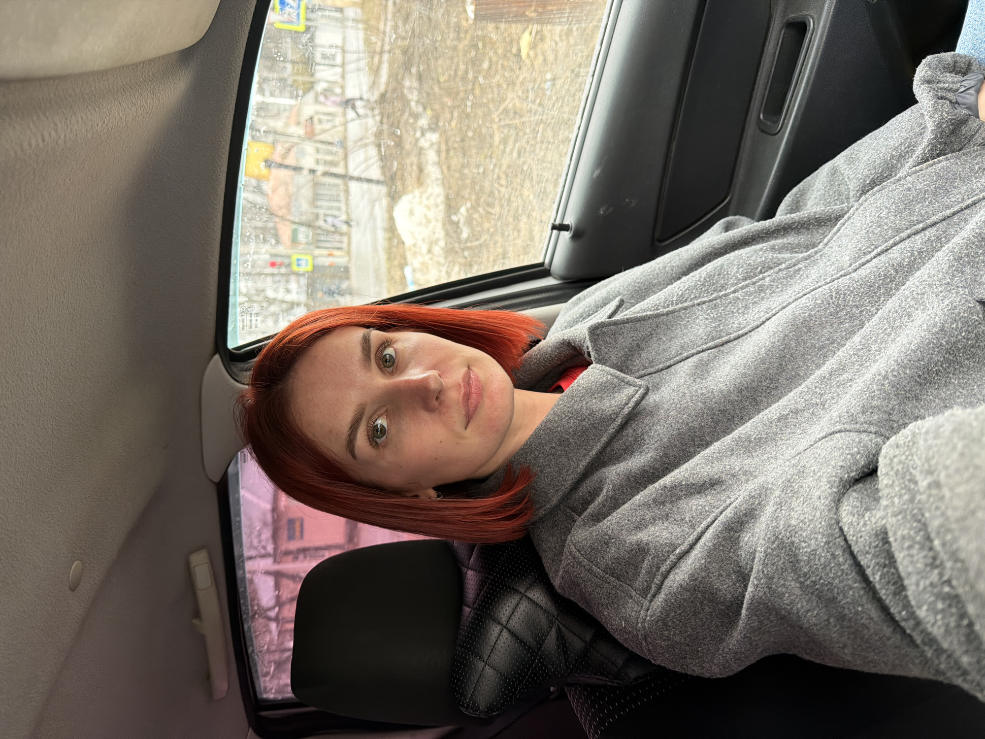 A woman with red hair is sitting inside a car, looking at the camera. She is wearing a gray coat. Through the car window, a street scene in Chaikovsky, Russia is visible, with wet roads, buildings, and street signs.