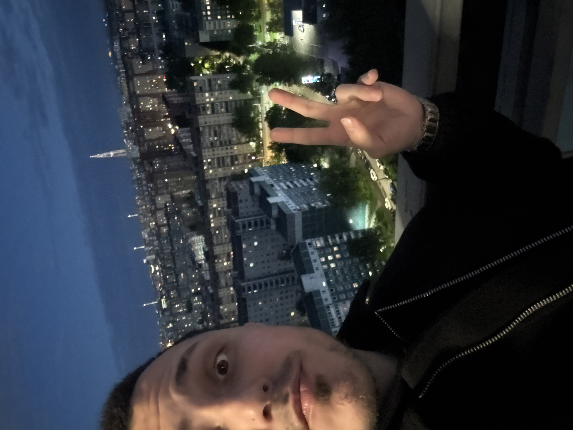 A selfie taken by a man is angled towards the left, capturing an urban skyline at twilight. The man, who is fair-skinned with dark hair and a goatee, is wearing a dark-colored jacket and a watch on his left wrist. He is holding up his hand in a peace sign. The background reveals a cityscape with numerous buildings lit up in the evening. A few green patches may be parks or treed areas. Based on the architecture and a distinctive tall spire in the distance, the location appears to be Saint Petersburg, Russia.