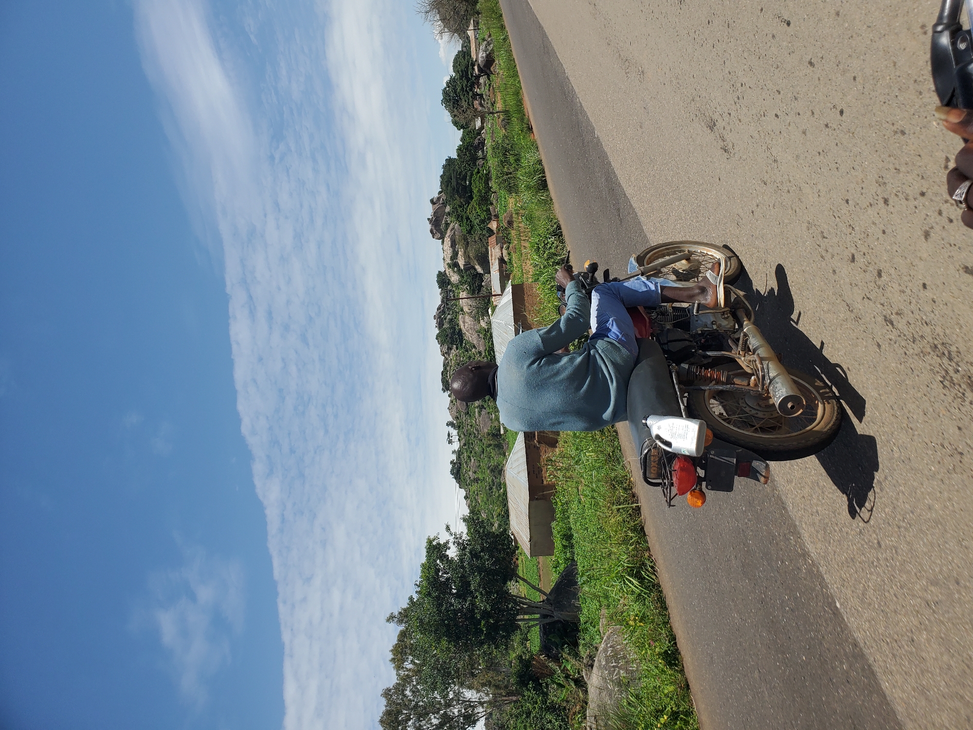 A person riding a motorcycle travels on a paved road through a rural area, possibly Panyam, Nigeria. The rider, seen from behind, wears a blue long-sleeved shirt and blue jeans. Their feet rest on the motorcycle's footpegs. The motorcycle is red and has a black seat and a white bottle attached to the rear. A shadow of the motorcycle and rider is cast on the road. The landscape features lush green vegetation, scattered trees, and rocky hills in the background. Several small structures with corrugated metal roofs are visible amidst the greenery, indicating a settlement. The sky is a bright blue with scattered white clouds.
