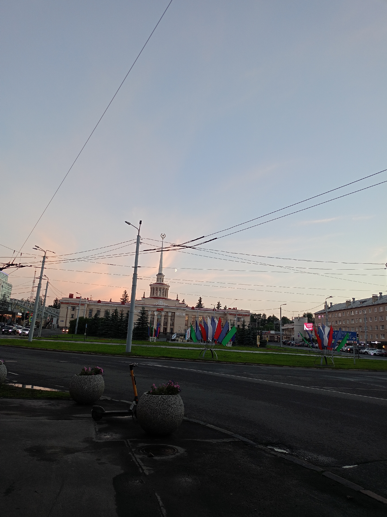 In the city of Unknown, Unknown, a wide, paved road occupies the foreground, with a slight sheen indicating recent moisture. To the left of the road, a sidewalk is visible, featuring two large, cylindrical planters, each containing small pink flowers. A dark-colored electric scooter is parked between these planters. The road continues into the background, bordered by a strip of green grass. Beyond the grass, a large, pale building with a central tower topped by a star stands prominently. This building is adorned with numerous small flags of various colors, including red, white, blue, and green, which are displayed in clusters. Further to the right, another building, a multi-story structure with numerous windows, is visible. Overhead, a complex network of power lines crisscrosses the sky, which transitions from a pale blue at the top to a soft, warm hue closer to the horizon, suggesting either dawn or dusk. A faint crescent moon is visible in the sky above the central tower. In the middle ground, several evergreen trees line the area in front of the pale building, and other buildings and traffic lights are present. Vehicles are seen on the road in the distance.
