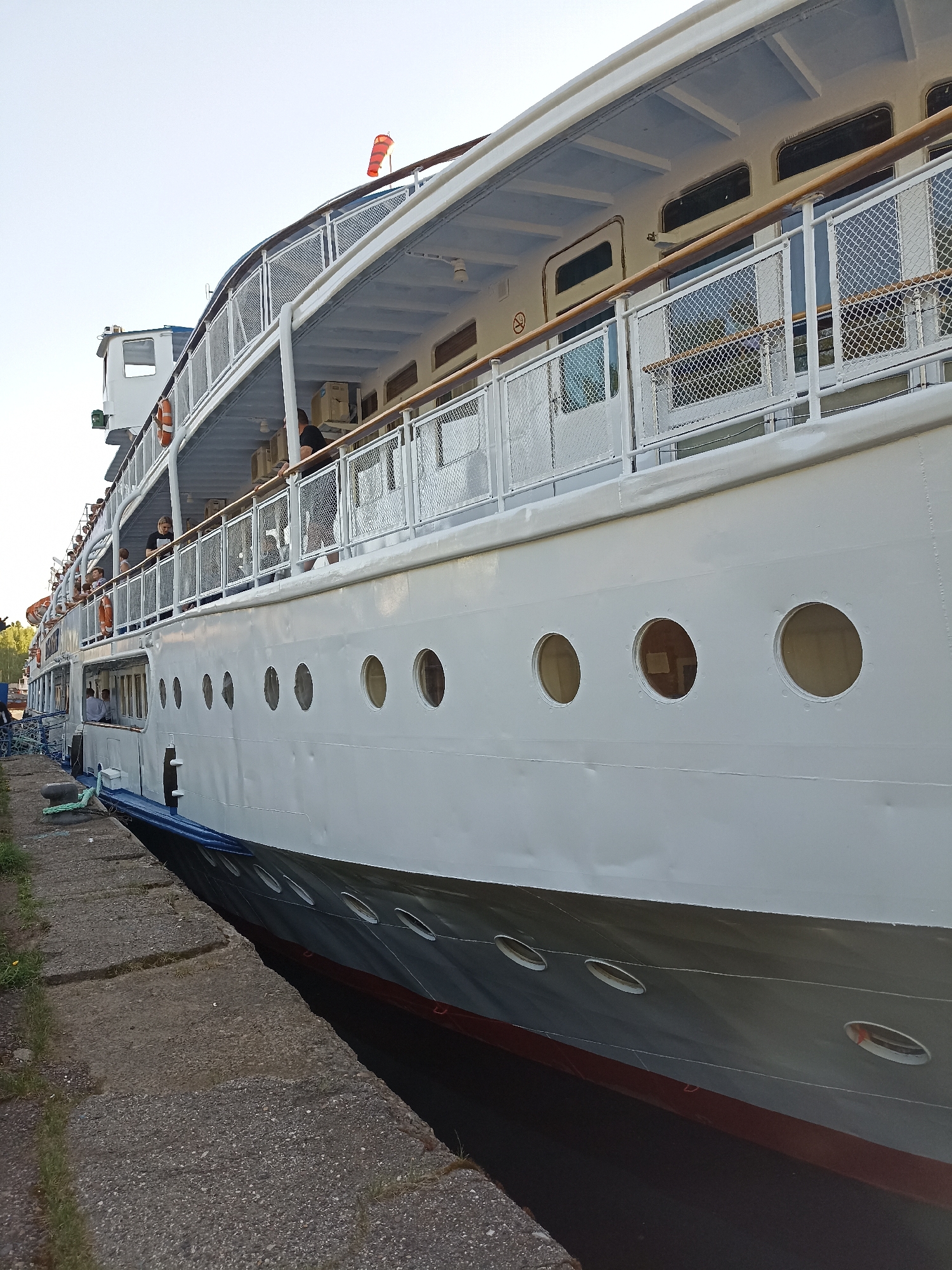 The image shows a large white river cruise ship docked next to a concrete embankment. The ship features multiple decks with windows and railings. Several people are visible on the deck of the ship, some standing by the railing. The hull of the ship has a series of circular portholes. The bottom of the ship's hull is gray with a red waterline stripe. The concrete embankment is cracked and has some grass growing along the edge. The location is unspecified, and the city and region are listed as "Unknown, Unknown." The sky is visible at the top of the frame, suggesting it is daytime.