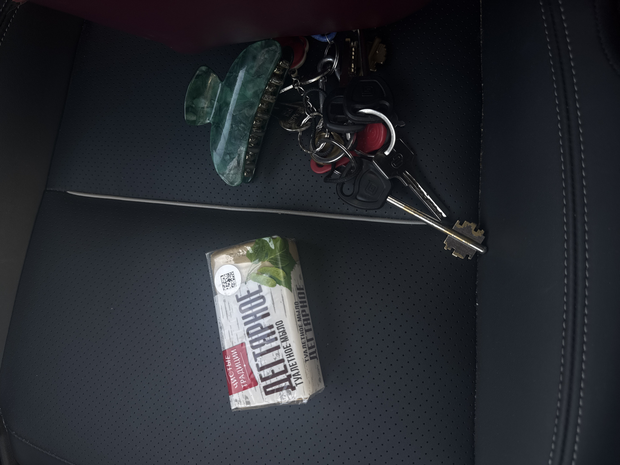 The image shows a close-up, high-angle shot of a black perforated car seat. On the seat are several items: a bar of "Detyarnoe" soap with Russian text and an image of birch leaves, a green plastic hair clip, and a set of keys on a keyring. The soap is in its packaging and is placed to the left of the hair clip and keys. The keys are a mix of silver and black, with some red key covers. The hair clip is positioned above and slightly to the right of the keys. The lighting appears to be natural, casting soft shadows. The image is cropped to focus on these objects and the texture of the car seat. The location of Chaikovsky, Russia, is not visually indicated in the image.