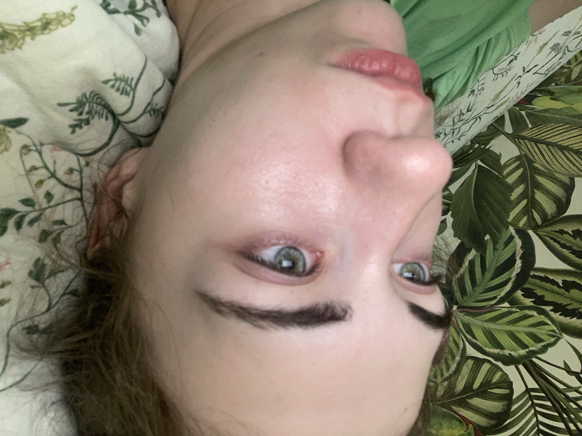 The image shows a close-up, side profile of a young Caucasian woman lying in bed. Her head is turned to her left, with her right eye and the right side of her face visible. Her eye is green and appears slightly puffy. Her eyebrows are dark and well-defined. Her lips are pink and slightly parted. She is wearing a green t-shirt. The background consists of bedding with a floral pattern and a wallpaper with large green leaves. The lighting is soft and diffused, suggesting an indoor setting. The image does not provide any information about the location in Zavodoukovsk, Russia, or any other geographical context.