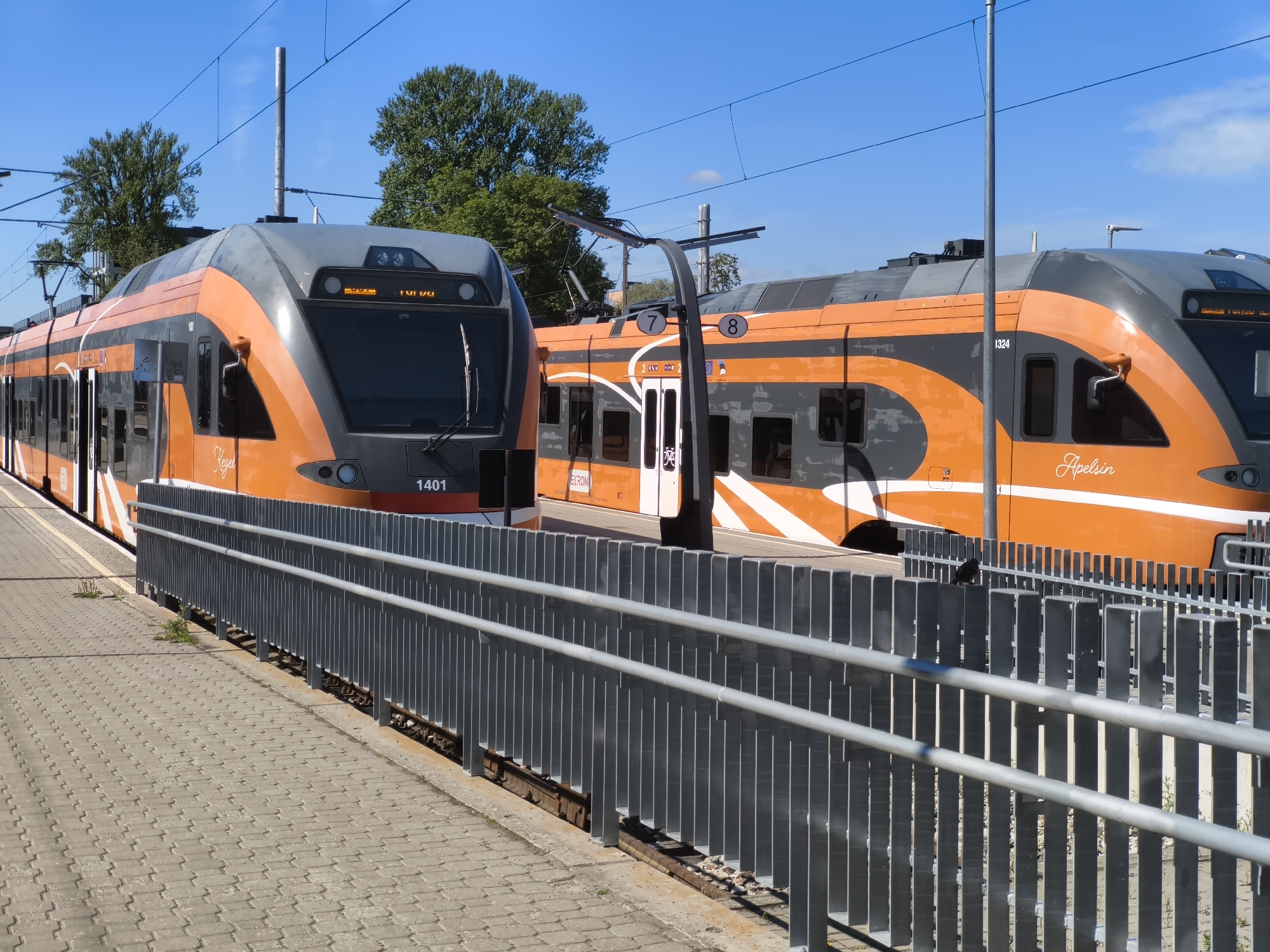 Two orange and grey electric trains are parked side-by-side at a train station platform in Tallinn, Estonia. The train on the left is closer to the camera and has the number "1401" visible on its front. The train on the right has numbers "7" and "8" on its side and the word "Apelsin" written on its front section. The platform is paved with grey interlocking blocks and is separated from the tracks by a modern grey metal fence. Overhead power lines are visible against a clear blue sky with scattered clouds. Trees can be seen in the background behind the trains.