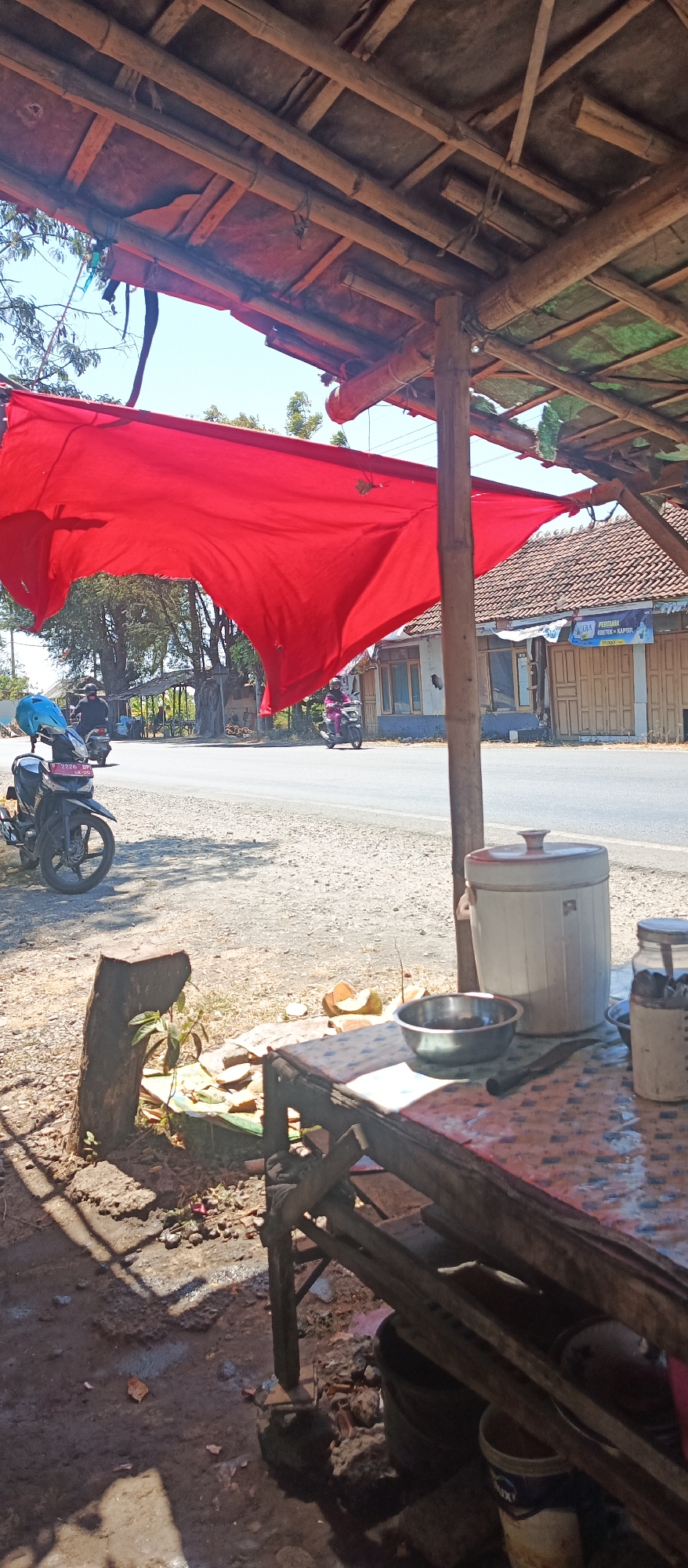 The image depicts an outdoor scene in Jatisari, Indonesia, under a rustic bamboo and fabric canopy. A large, vibrant red fabric drapes prominently in the foreground, partially obscuring a glimpse of a roadside scene. A paved road is visible, with two motorcycles passing by. One motorcycle, a black scooter with a blue helmet on its handlebars, is parked on the left side of the road. Another motorcycle, ridden by a person wearing a pink outfit, is seen further down the road.
In the immediate foreground, under the canopy, a wooden table is set with various items. A large, white cylindrical container with a lid stands on the ground to the right of the table. On the table itself, a metal bowl, a knife, and a glass jar filled with utensils are present. A half-cut coconut sits on the table, and scattered coconut pieces are on the ground. A tree stump with a small plant growing from it is located to the left of the table. The ground is a mixture of dirt and gravel. In the background, houses with tiled roofs and closed wooden doors line the roadside. The sky is clear and bright blue, suggesting a sunny day.