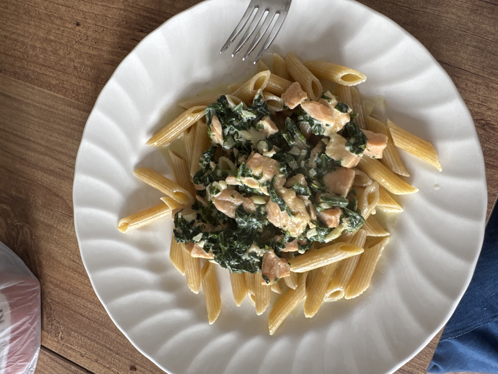 The image shows a top-down view of a plate of penne pasta topped with a mixture of spinach and salmon in a creamy sauce. A fork is partially visible in the upper center of the plate. The plate is white with a scalloped edge and rests on a wooden surface. A corner of what appears to be a plastic bag is visible in the lower left corner, and a blue garment is visible at the lower right edge of the image. The location and city are unknown.