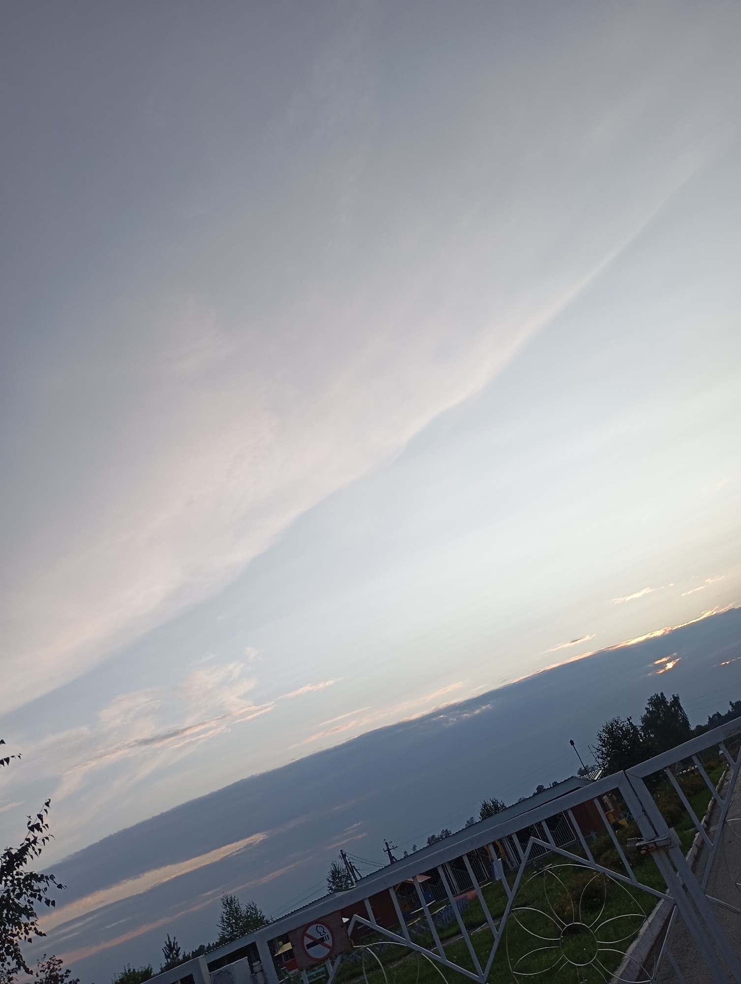 The image depicts an outdoor scene, likely a park or residential area, in the city of Фоки, Russia. The composition is dominated by a vast sky with varied cloud formations. The sky transitions from a pale blue or grey at the top left to a brighter, lighter hue towards the right, suggesting the presence of sunlight, possibly near sunset or sunrise, as indicated by subtle orange and yellow tones peeking through some clouds near the horizon.
In the lower portion of the image, a metal fence with a decorative floral pattern is visible. Beyond the fence, there are glimpses of buildings with pitched roofs, suggesting structures like houses or a community center. Some greenery, including trees and grass, is present, interspersed with the man-made structures. A "no smoking" sign is attached to the fence, indicating a designated non-smoking area. The overall impression is of a calm, possibly late afternoon or early evening setting in a suburban or semi-rural environment. The image is tilted, giving a dynamic perspective to the sky and the landscape.