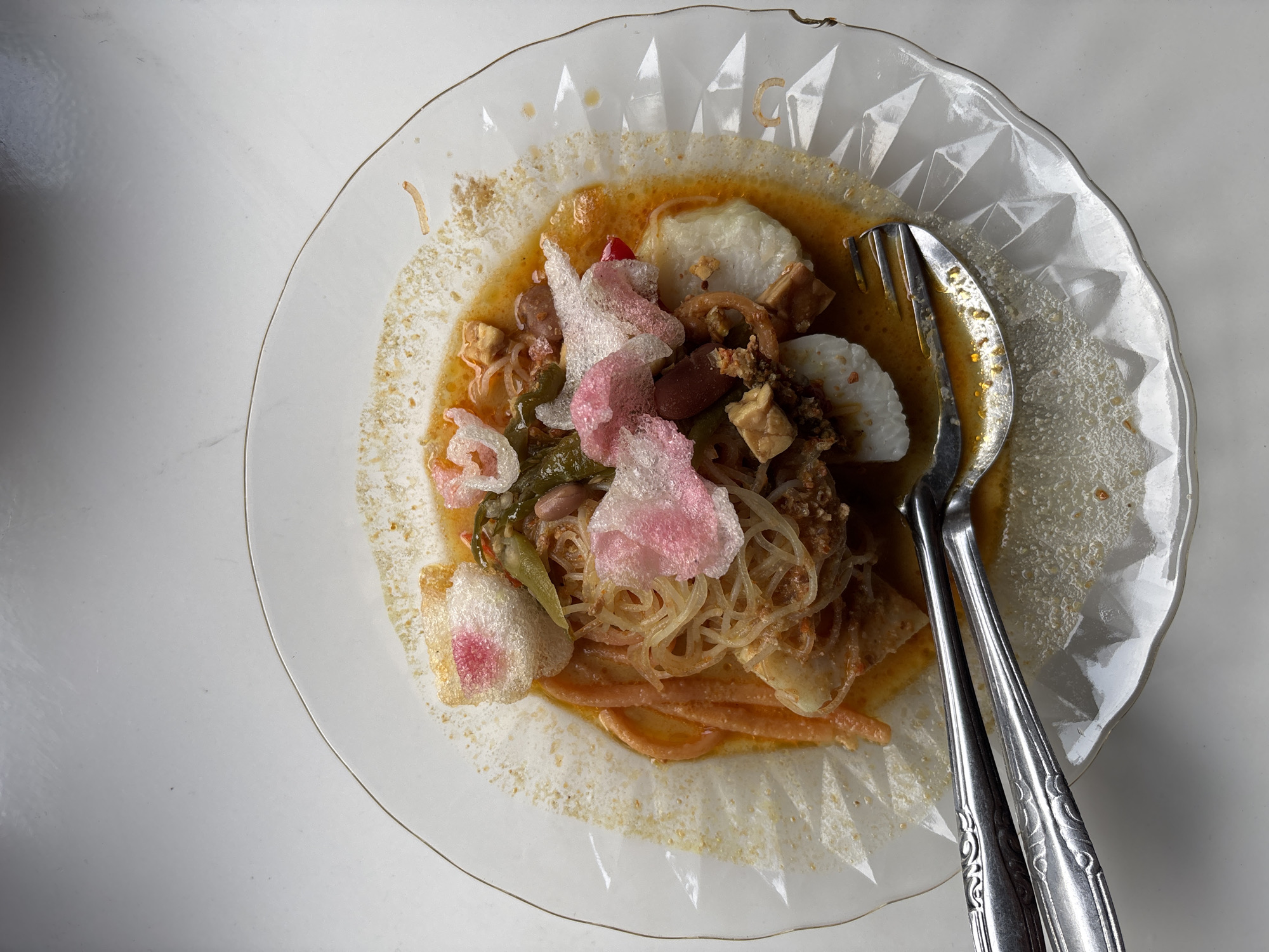 This overhead shot captures a plate of an Indonesian savory dish, likely Lontong Sayur or a similar meal, served on a clear, scalloped glass plate with a diamond-patterned rim. The dish is rich with various ingredients submerged in a yellowish-orange sauce or gravy.
Central to the meal are white, oval-shaped chunks, which appear to be lontong (compressed rice cakes). Intermingled with these are fine vermicelli noodles, small cubes of tofu or tempeh, green vegetables (possibly long beans), and reddish-brown beans. Adding a vibrant, crunchy element are several pieces of puffed crackers (krupuk), some white and others a striking pink, scattered on top.
A silver-colored metal fork and spoon, both featuring decorative patterns on their handles, rest within the food on the right side of the plate, indicating the meal is ready to be eaten. The plate is positioned on a clean, smooth, light-colored surface, suggesting an indoor dining setting. The overall lighting is bright and even, characteristic of an interior during daytime or under artificial lights. This type of cuisine is culturally significant in Indonesia, aligning with the location context of Sangso, Indonesia.