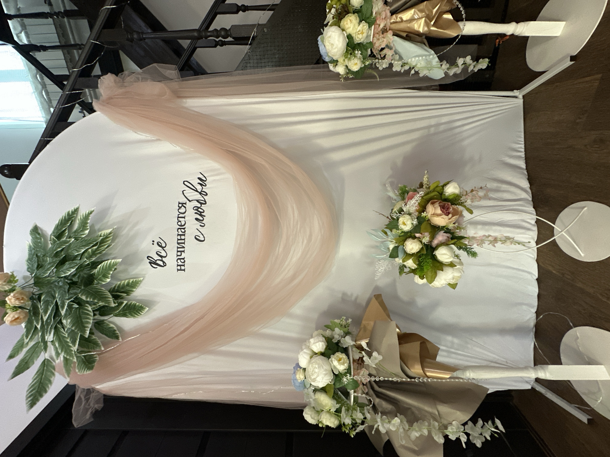 Here is a detailed description of the image:
The image shows a wedding decoration setup. The main element is a white, semi-circular structure with peach-colored draping on the left side. The phrase "Всё начинается с любви" (Everything starts with love) is written on the white surface. Floral arrangements are placed on the top corners and lower right corner. The structure is supported by white stands with circular bases. The setup is indoors, with wooden stairs in the background.