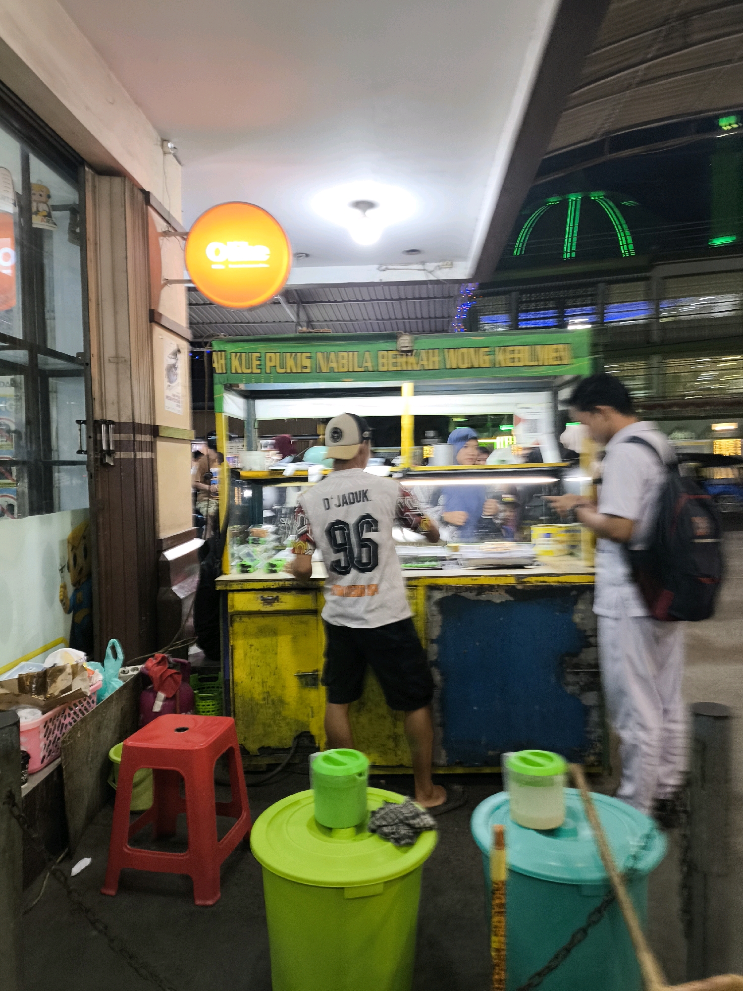 The image depicts an outdoor night scene at a food stall in Gorontalo, Indonesia. The stall has a green awning with Indonesian text. In the foreground, there is a red plastic stool. To the right, a man wearing a white t-shirt with the number "96" and a cap is standing and looking towards the stall. He is facing away from the camera. To his right, another person, possibly a man, wearing white pants and a white shirt, with a black backpack, is standing and looking at a phone. Inside the stall, there are items on display, and a person wearing a blue head covering is visible behind the counter. A round, orange sign with the word "Ofline" is mounted on the wall to the left. The overall lighting suggests a commercial area at night.