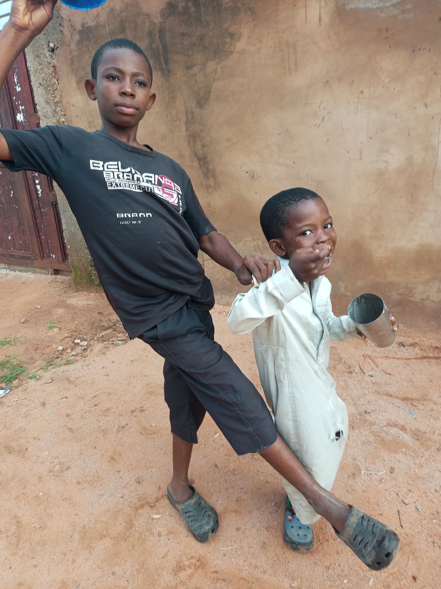 In Tempure, Nigeria, two young boys stand on a dirt ground in front of a textured, tan wall. The boy on the left, wearing a black t-shirt with white lettering that reads "BELL BRAND EXTREME CUT," and dark pants, is holding a blue object above his head. The boy on the right, dressed in a light-colored button-down shirt and pants, is smiling and holding a metal cup in his right hand, while his left hand is near his mouth. A third person's hand is visible on the right boy's left shoulder. The ground is uneven with scattered pebbles and sparse dry grass. A weathered wooden door is visible on the left side of the image.