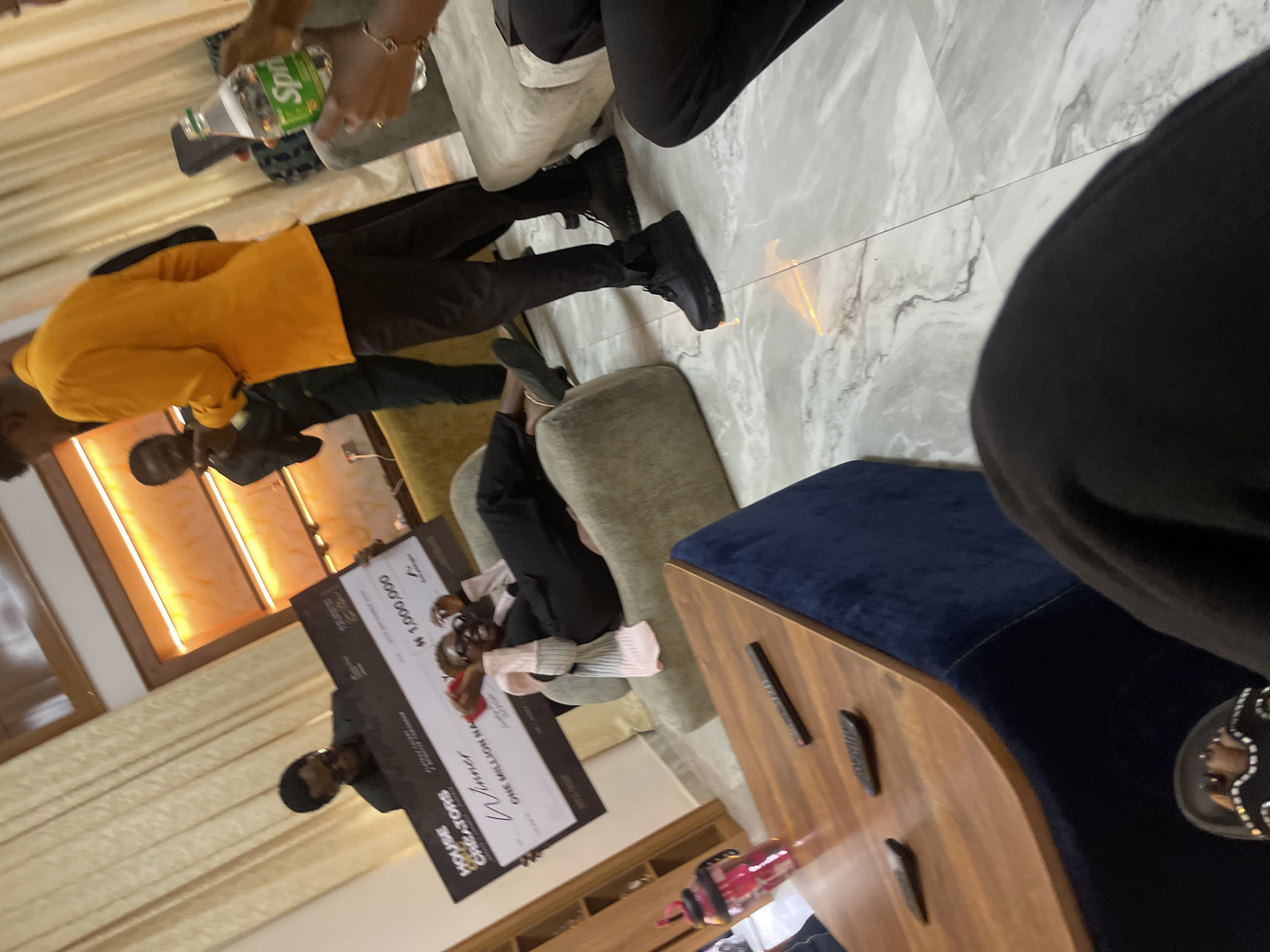 The image depicts an indoor scene at what is identified as the "House of Creators" in Gura Top, Nigeria. The central focus is a large novelty check with the words "ONE MILLION NAIRA" and "# 1,000,000" printed on it, indicating a prize or award. A woman, dressed in black and wearing glasses and a pink scarf, is seated on a light-colored couch, smiling and holding her hands up in excitement as she poses with the check.
Several individuals are present in the frame. To the left of the woman, a man in a dark suit and glasses is seen holding up the check. Another man, wearing an orange blazer, stands to the right of the woman, facing away from the camera, with his left hand extended. Partially visible in the background are other people and elements of the room's decor, including large curtains, illuminated wall panels, and other seating.
In the foreground, a wooden coffee table with a dark blue upholstered base is visible. It holds several remote controls and a pink water bottle with a straw. The flooring appears to be marble or a similar polished material. The overall atmosphere suggests a celebratory event or announcement.