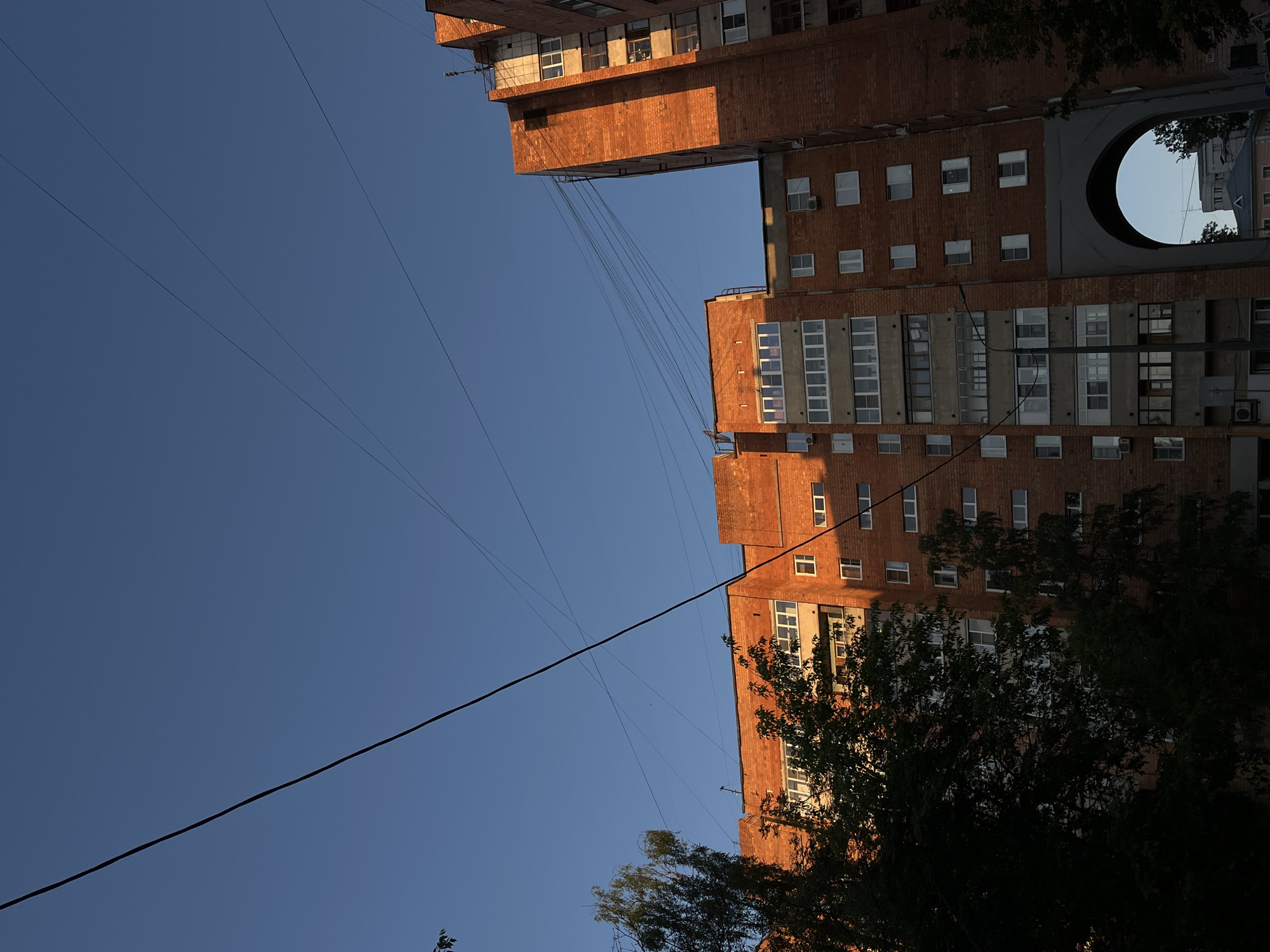 The image shows a brick apartment building in the city of Nizhny Novgorod, Russia, bathed in the warm light of the setting sun. The building features multiple stories with numerous windows and balconies. A large archway is visible on the lower section of the building. Overhead, electrical wires crisscross the clear blue sky. Dark green trees are silhouetted against the building in the foreground. The overall scene depicts a residential area in the city during the golden hour.