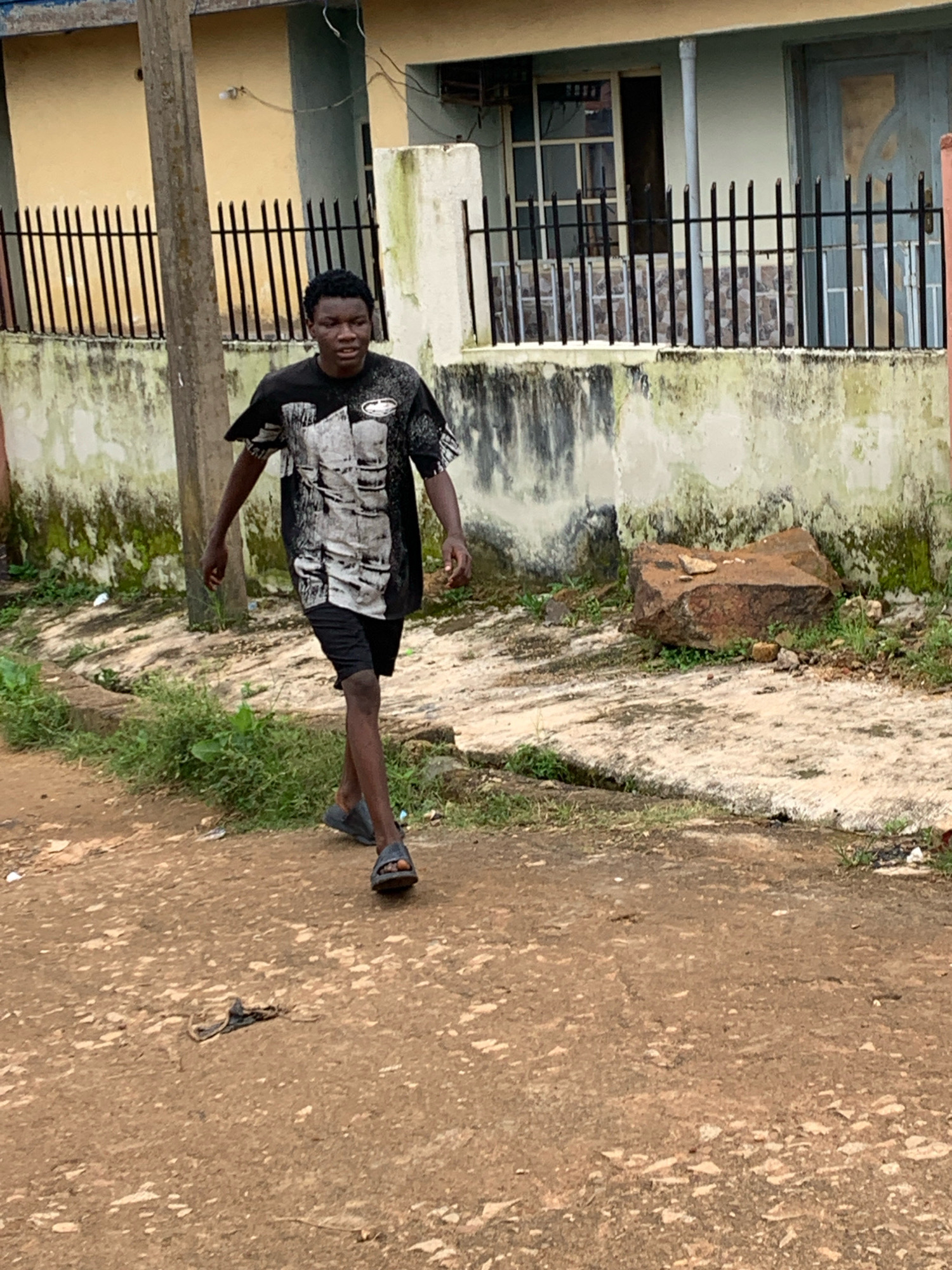 A young Black male with short, dark, curly hair walks towards the camera on a dirt road. He is wearing a black t-shirt with a graphic design in white and grey, black shorts, and black sandals. The background features a light-colored building with a metal fence in front of it. The building has green and white staining on its exterior walls, indicating potential water damage or age. To the right of the building, a large, reddish-brown rock sits on the ground. Green vegetation is visible along the edge of the road and near the building. The overall environment appears to be an outdoor urban setting, possibly in Jos, Nigeria, given the architectural style and the presence of dirt roads and lush vegetation. The ground is uneven and dusty, with some scattered debris.