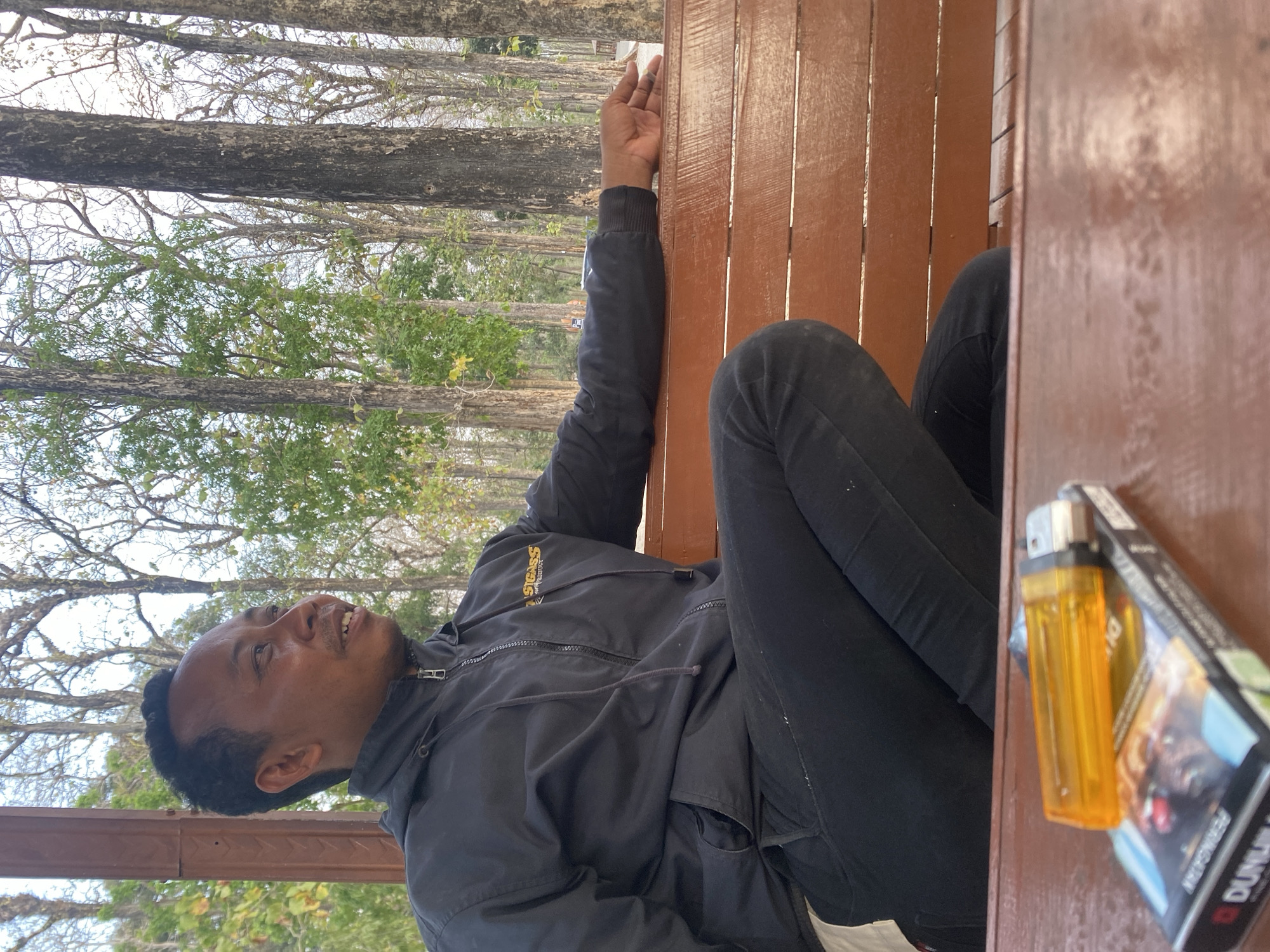 A man sits on a wooden bench outdoors, with trees in the background. He is wearing a black hooded jacket with a zipper and black pants. A pack of cigarettes and a lighter are on a wooden table in the foreground. The man is looking to his right and his left arm is resting on the bench.