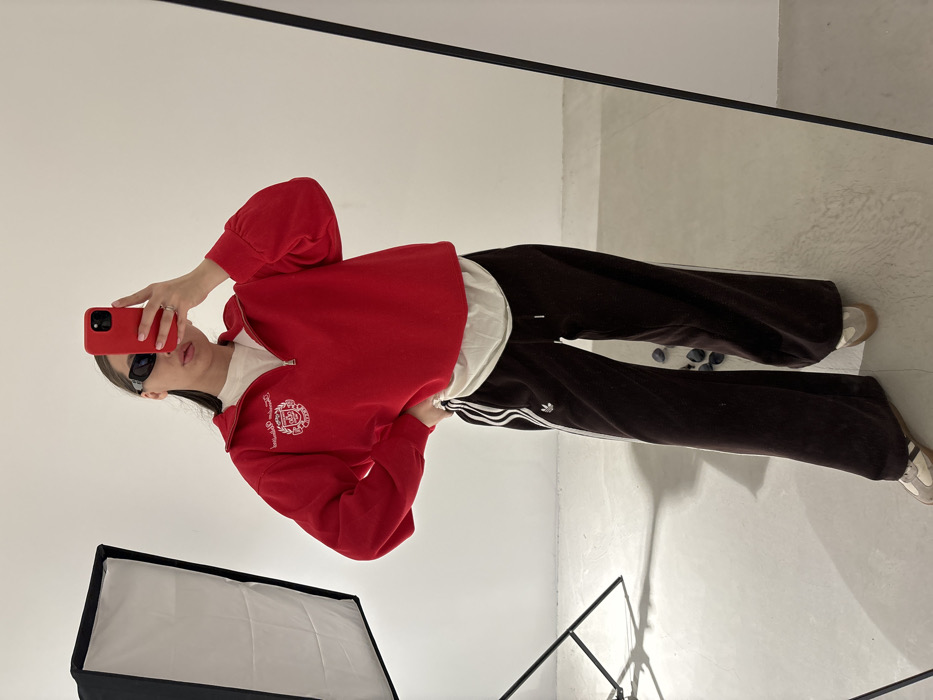 A young adult female is depicted standing in what appears to be an indoor studio or well-lit room, captured via a full-length mirror selfie. She holds a red-cased smartphone in her right hand, obscuring her upper face, while her left hand rests on her hip. The individual wears dark rectangular sunglasses, a red long-sleeved quarter-zip sweatshirt with a white embroidered crest on the left chest, layered over a light-colored t-shirt. She is dressed in dark brown, wide-leg athletic pants featuring white side stripes and an Adidas trefoil logo on the left thigh, complemented by light-colored athletic shoes. A ring is visible on her right hand's ring finger. To the left of the subject, a black photography softbox on a stand is present. The background consists of uniform white walls and a light-colored floor, on which several small, dark, irregular objects are scattered near the subject's feet. The specific location within Krasnodar, Russia, cannot be visually identified from the image.