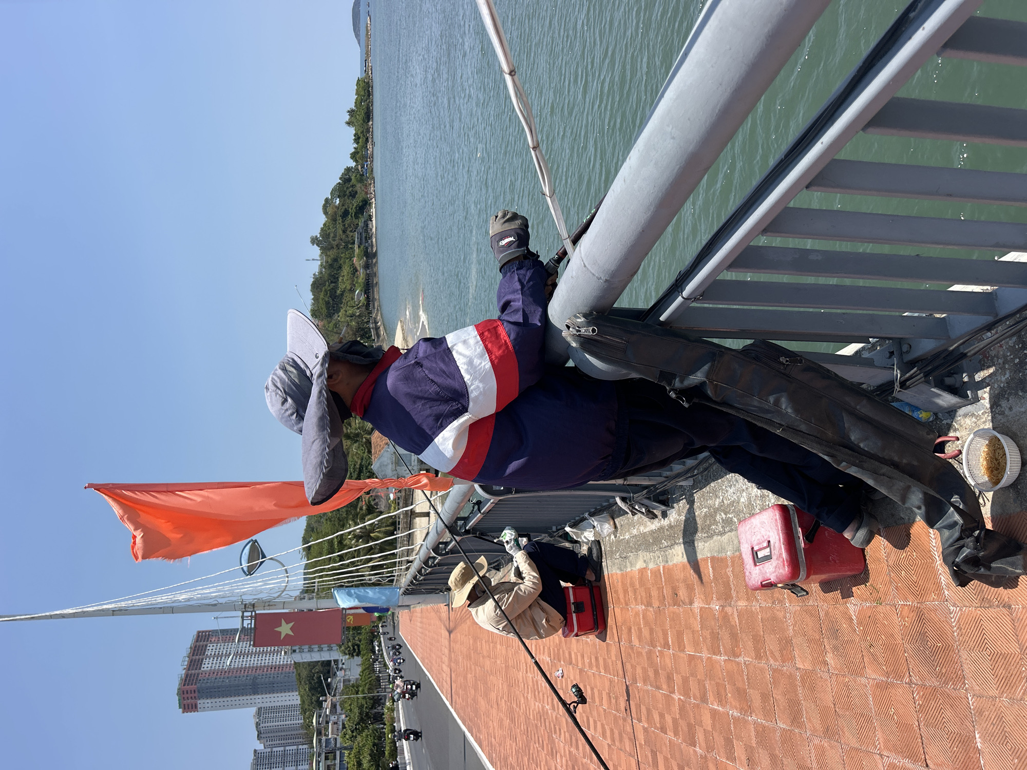 The image captures two men fishing from a bridge in Nha Trang, Vietnam, on a clear, sunny day. The bridge features a distinctive reddish-brown tiled walkway.
In the foreground, a man wearing a blue jacket with red and white stripes and a gray sun hat is seen leaning against a grey metal railing. He is actively engaged in fishing, holding a rod that extends over the shimmering body of water below. Beside him, a black fishing bag and a white bowl containing what appears to be bait or snacks are visible on the bridge floor. Further along the bridge, another individual, dressed in a beige jacket and a straw hat, is seated on a red cooler, also fishing with a rod extending towards the water.
The background features a wide expanse of water, likely the ocean or a large river, bordered by lush, green, tree-covered hills. In the distance, a cityscape with tall modern buildings is visible. Overhead, the bridge's intricate cable-stayed structure is prominent. A vibrant orange-red flag and the national flag of Vietnam (red with a yellow star) are flying from poles attached to the bridge cables, affirming the location. The sky is bright blue and cloudless, indicating excellent weather conditions. The scene conveys a sense of peaceful leisure and focused activity.