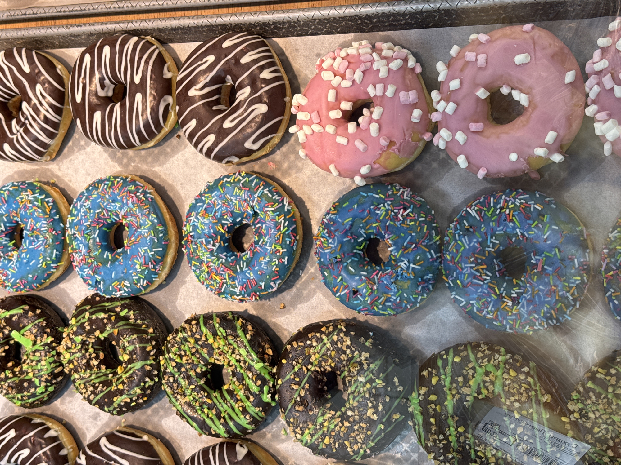 An overhead shot captures a vibrant display of various donuts arranged in rows, likely in a bakery or supermarket case. The donuts are presented on a light-colored liner, possibly parchment paper, within a metallic tray.
The top row features dark chocolate-glazed donuts with white icing drizzles on the left, and pink-iced donuts topped with small white and pink marshmallow pieces on the right. One of the pink donuts shows slight damage to its icing.
The middle row consists entirely of bright blue-iced donuts generously covered with a mix of colorful sprinkles, including red, yellow, blue, white, and green.
The bottom row displays dark chocolate-glazed donuts adorned with green icing drizzles and a scattering of crushed nuts or seeds. One of these bottom-row donuts has a small white rectangular sticker with black text partially visible. While most of the text is obscured or distorted by reflections, the numbers "53.03" and some Cyrillic letters like "CAX96" are faintly discernible, suggesting a price or product label.
The lighting is bright, illuminating the various toppings and glazes. The overall impression is one of a colorful and enticing selection of sweet treats. Slight reflections on what appears to be a protective barrier, like glass or plastic, are visible across the image.