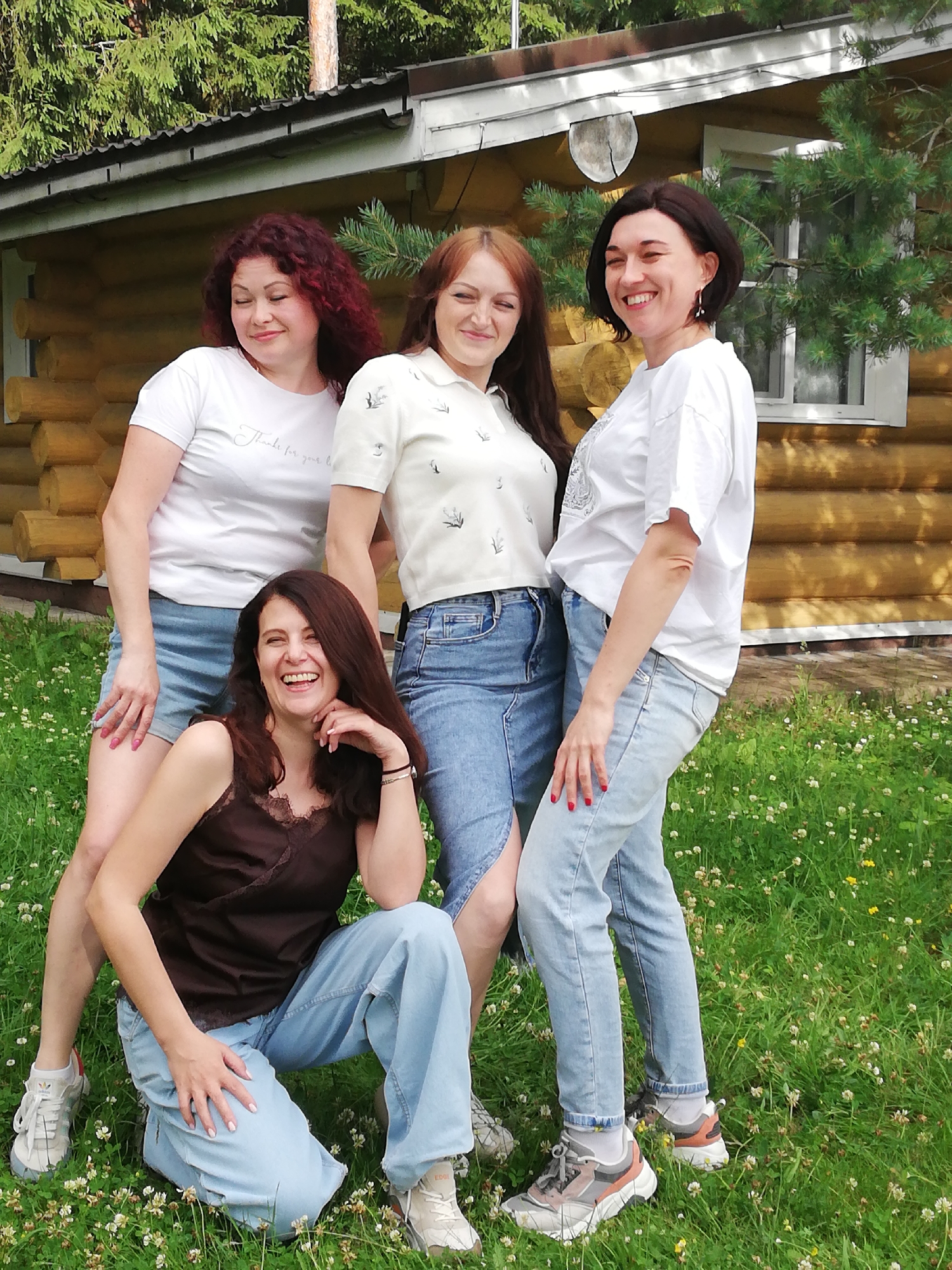 Four women are posing in front of a wooden cabin surrounded by lush green grass and trees. The woman on the far left has long, curly red hair and is wearing a white t-shirt and blue denim shorts. Next to her, a woman with long brown hair smiles broadly, wearing a white collared shirt with an embroidered pattern and a denim skirt. The third woman from the left has short dark hair and is wearing a white t-shirt and light-wash jeans. She is laughing with her head tilted back. The woman in the foreground is kneeling on the grass, looking up and laughing with her hand on her chin. She has long dark brown hair and is wearing a dark brown camisole and light-wash jeans. The cabin appears to be made of natural wood logs and has a dark roof. The scene is set outdoors on a sunny day. The overall atmosphere is casual and friendly. The location of Шапки, Russia, is not explicitly indicated in the image, but the wooden cabin and surrounding vegetation suggest a rural or natural setting.