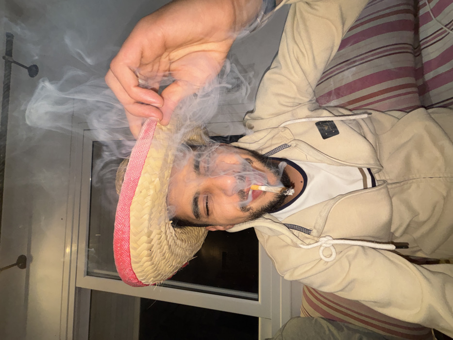 This image depicts an indoor scene, consistent with a private residence, in what is indicated to be Martil, Morocco. The central subject is an adult male, likely in his 20s or 30s, with dark hair and a beard, looking directly at the camera with his left eye closed and his right eye partially open. He is wearing a beige zippered hooded jacket over a white t-shirt with brown stripes, and a light-colored straw hat with a red brim. His right hand is raised, gripping the brim of the hat as if adjusting it. A lit cigarette is positioned between his lips, actively burning and emitting a significant plume of smoke that partially obscures his face and rises upwards. The smoke is prominent around his mouth and nose. He is seated on a sofa or cushioned bench with red and white striped upholstery. In the background, a large window or glass door with dark panes is visible, suggesting nighttime or an unlit exterior. Above, two ceiling-mounted light fixtures or similar hardware are discernible against a light-colored ceiling.