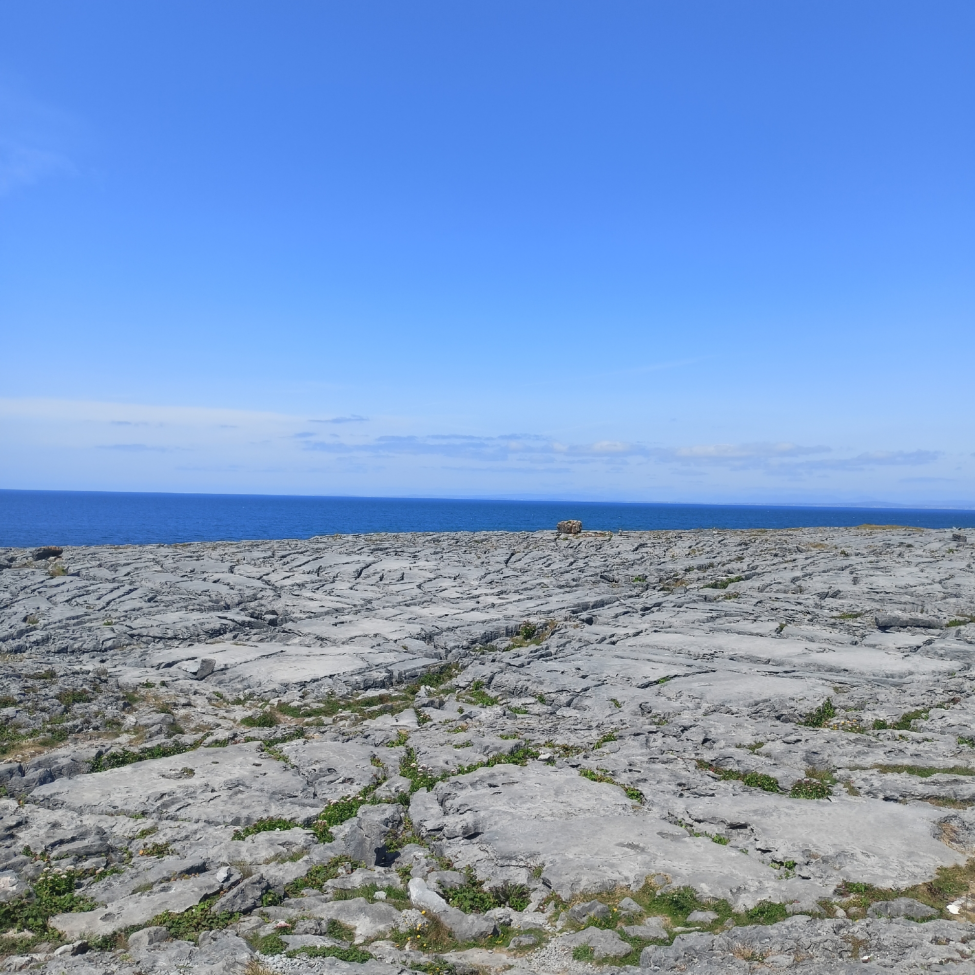 The image shows a vast, grey, rocky landscape with a bright blue sky and the ocean in the background. The rocks are flat and fractured, with small patches of green vegetation growing in the crevices. The ocean is a deep blue, meeting the horizon under a sky with a few wispy clouds. In the middle distance, a small, rough-hewn stone structure or cairn sits on the rocks. The scene appears to be a natural coastal environment, possibly the Burren region of County Clare, Ireland, known for its limestone karst landscape. No people are visible in the image.
