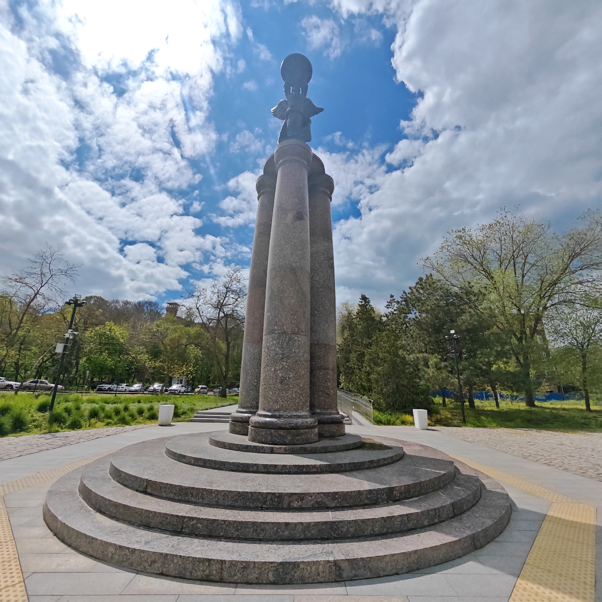 The image shows a monument in Taganrog, Russia. The monument consists of a statue on top of three granite columns, which are on top of a multi-tiered circular base made of the same granite. The statue appears to depict a figure holding a sphere or globe. The monument is located in a park-like setting with trees and grass. In the background, there is a building with a tower visible through the trees. Cars are parked on a street nearby. The sky is partly cloudy. The monument and its surrounding area look like a tourist location.