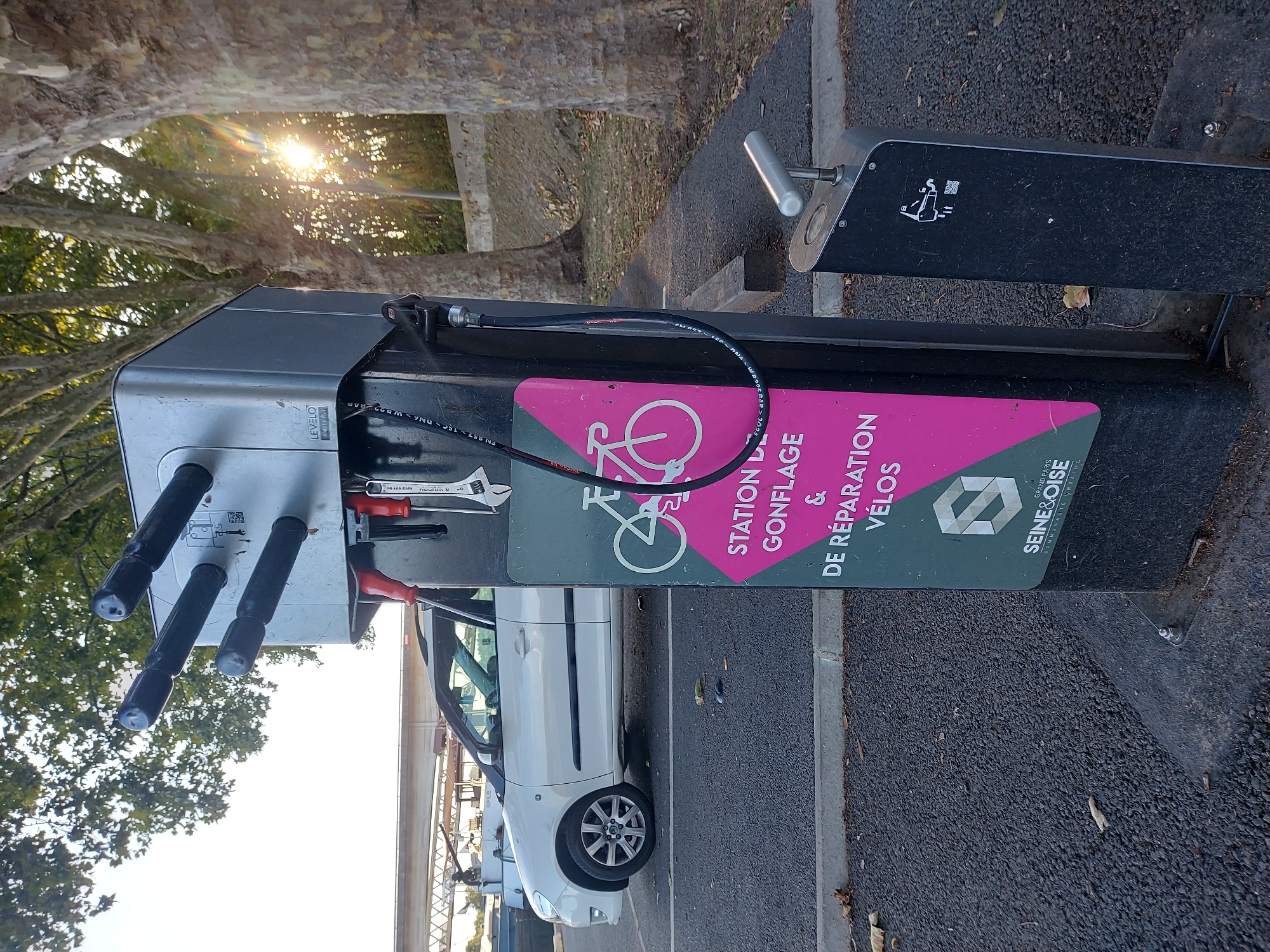 The image depicts a bicycle repair and inflation station located in Conflans-Sainte-Honorine, France. The station is a metallic structure with several nozzles for tire inflation, and a selection of tools like wrenches are attached to its side. A hose is coiled and attached to the station. A sign on the station reads "STATION DE GONFLAGE & DE RÉPARATION VÉLOS," which translates to "TIRE INFLATION & BIKE REPAIR STATION." Below this text, the logo for "SEINE & OISE" is visible, indicating the administrative region or a local service. In the background, a white car is parked, and trees line a paved area. The sun is visible, casting a lens flare through the trees, suggesting it is either morning or late afternoon. The ground is asphalt.