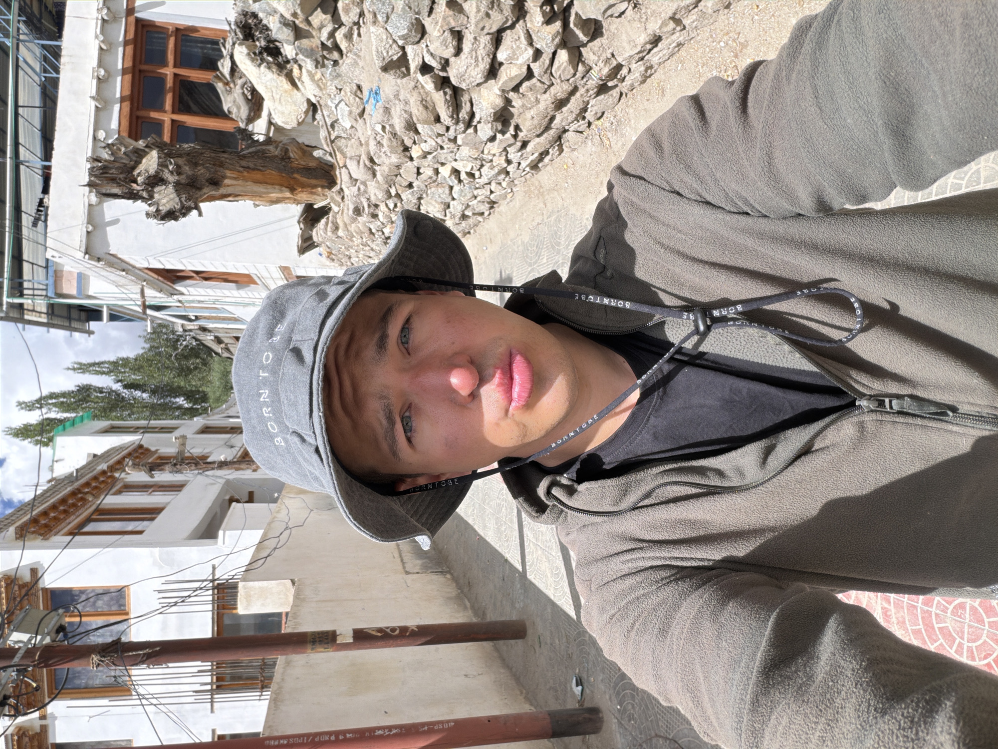A young man is taking a selfie outdoors in a narrow street in Leh, India. He is wearing a grey bucket hat with the text "BORN TO BE" visible on it. He has on a dark t-shirt and a grey fleece jacket with a zipper. A string with the text "BORN TO BE" is hanging from his hat. The street is paved with stone and has a wall made of stacked rocks on the right side. Buildings with white walls and wooden window frames are visible on both sides of the street. There are also utility poles with wires and some trees in the background. The lighting suggests it is daytime.