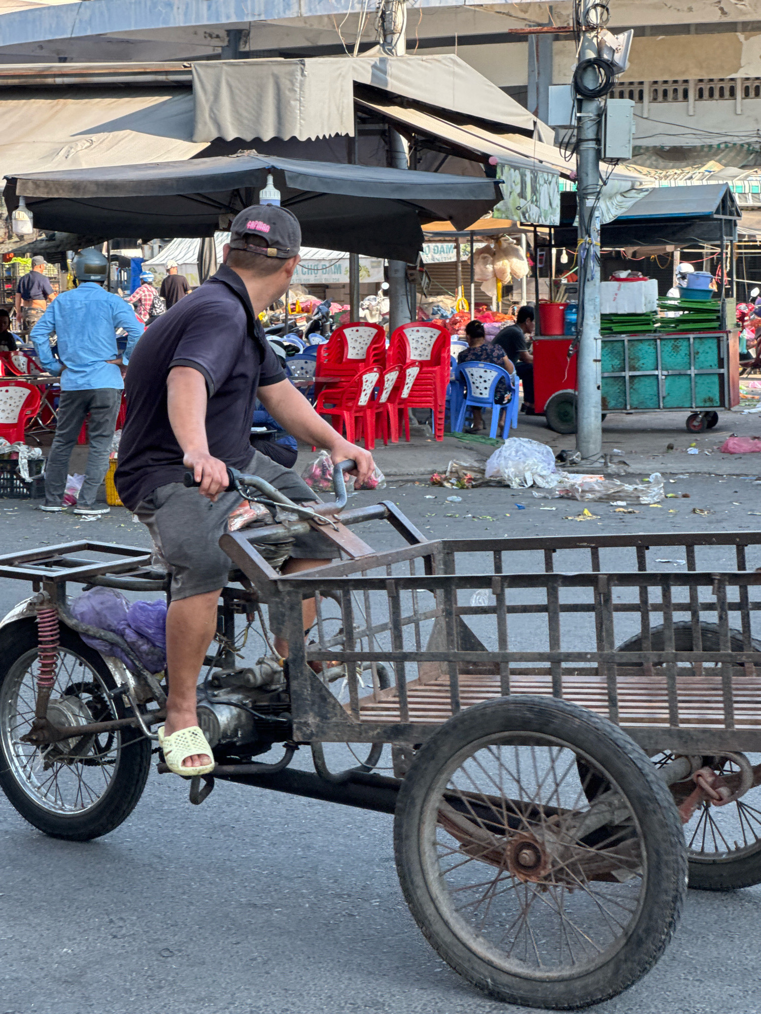 A man rides a three-wheeled motorcycle cart through a busy outdoor market in Nha Trang, Vietnam. The man wears a dark blue short-sleeved shirt, grey shorts, and yellow slides. The motorcycle cart is weathered and appears to be made of metal. It has a basket-like structure at the rear, presumably for carrying goods. The market environment is evident, with numerous stalls, awnings, and other people visible in the background. Several red plastic chairs are stacked near a stall, and various goods are displayed. Debris is scattered on the paved ground. The scene suggests a typical day in a Vietnamese urban marketplace, characterized by activity and commerce.