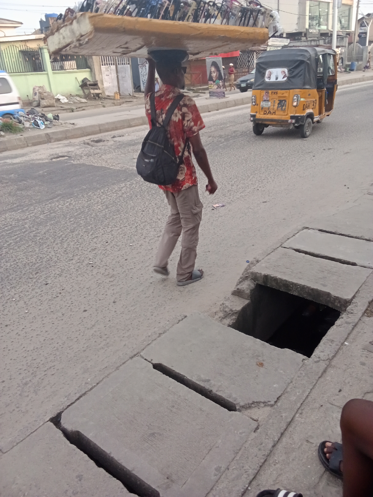 The image captures a bustling street scene in what appears to be an urban or semi-urban area. The central figure is a person, seen from behind, walking on the side of an asphalt road while expertly balancing a large, flat, rectangular object, possibly a mattress or foam, on their head. They wear a red and white patterned short-sleeved shirt, light brown pants, and light-colored sandals, with a black backpack slung over their shoulders.
To the right of the person, a concrete sidewalk features an exposed drainage system. Several concrete slabs cover parts of the ditch, but one section is wide open, revealing a deep, dark cavity below. In the bottom right corner, the legs and feet of another person, wearing black slides with white stripes, are visible, suggesting they are seated or crouching nearby, possibly the photographer.
In the middle ground, a yellow auto-rickshaw (often called a "Keke Napep" in Nigeria) is either parked or slowly moving on the road. The rear of the vehicle is visible, displaying "TVS" on its canopy and a license plate that reads "DAH 39", with "DAH" also visible below it. Further in the background, various buildings line the street, including one with a light green fence and another commercial building with the word "GOGGLES" partially visible on its facade. A large billboard featuring a woman's face stands near the road. The road itself shows signs of wear, with visible cracks and a textured surface. Scattered debris and litter are noticeable along the roadside, particularly near the green fence.
The lighting suggests it is daytime, possibly late afternoon, with no harsh shadows indicating midday sun. The overall impression is a vibrant street environment, consistent with a location like Ado, Nigeria.