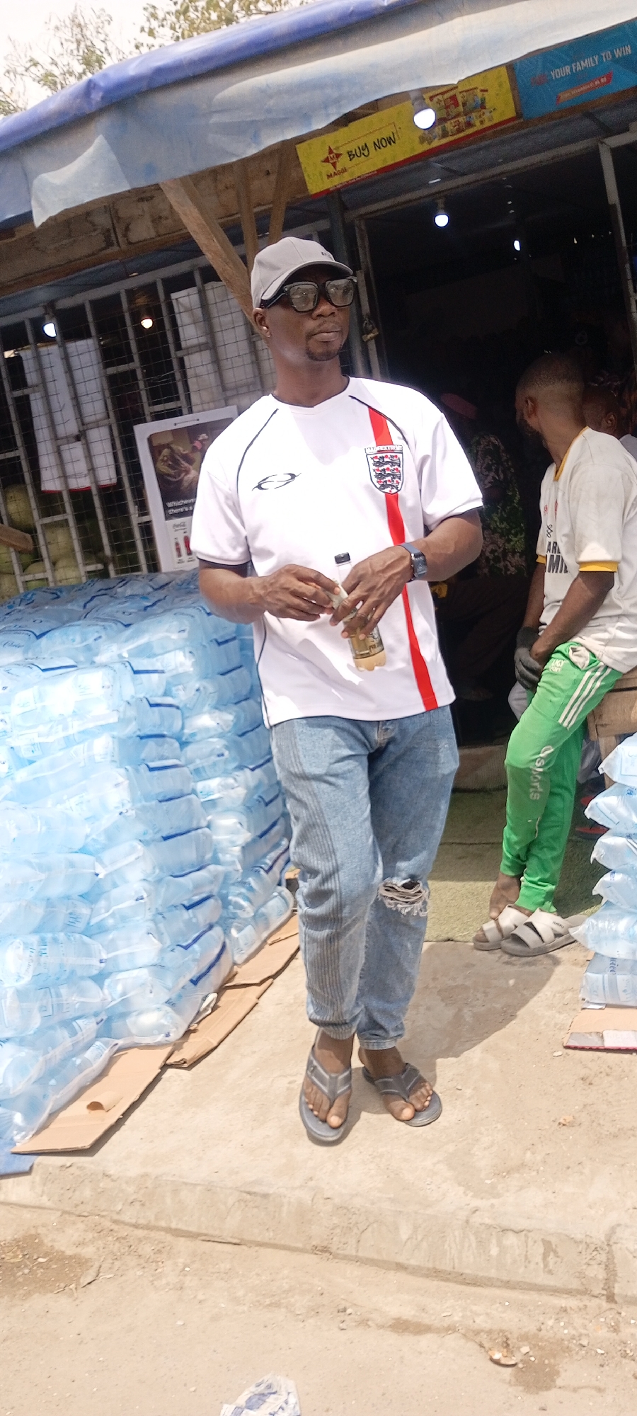 The image shows a man standing in front of what appears to be a retail establishment or market stall. The location can be identified as the city of Gayawa, Nigeria.
The man is wearing a white t-shirt with a red stripe on the side and an England football crest on the chest, light blue jeans with a rip at the knee, a gray baseball cap, and dark sunglasses. He is holding a small bottle in his hands.
To his left is a large stack of bottled water. In the background, there's another person wearing a white t-shirt and green pants, seated, with more stacks of bottled water nearby. There are signs above the stall, some featuring advertising and brand names. The stall appears to be constructed with a corrugated metal roof. The ground is a light-colored concrete or paved surface.
