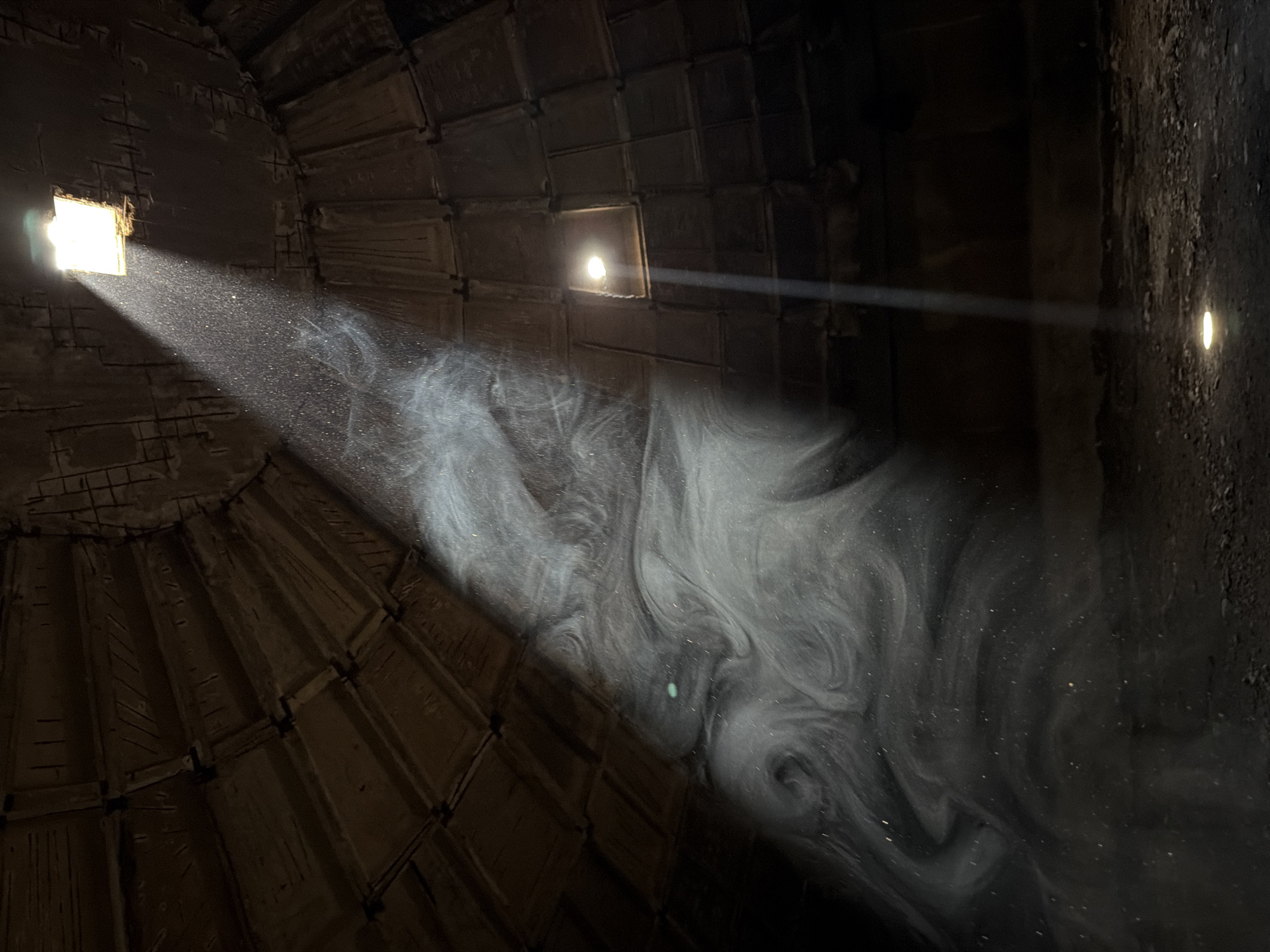 The photograph appears to be taken inside a dark, possibly concrete structure, likely an interior dome or enclosed space. Light is entering from two separate small square openings near the top, with dust particles visible in the beams of light. The concrete structure has a patterned texture, composed of rectangular blocks. The light casts a spotlight on the dusty environment, creating a swirling pattern in the center. There are beams of light appearing on the upper right. The location is Tbilisi, Georgia.