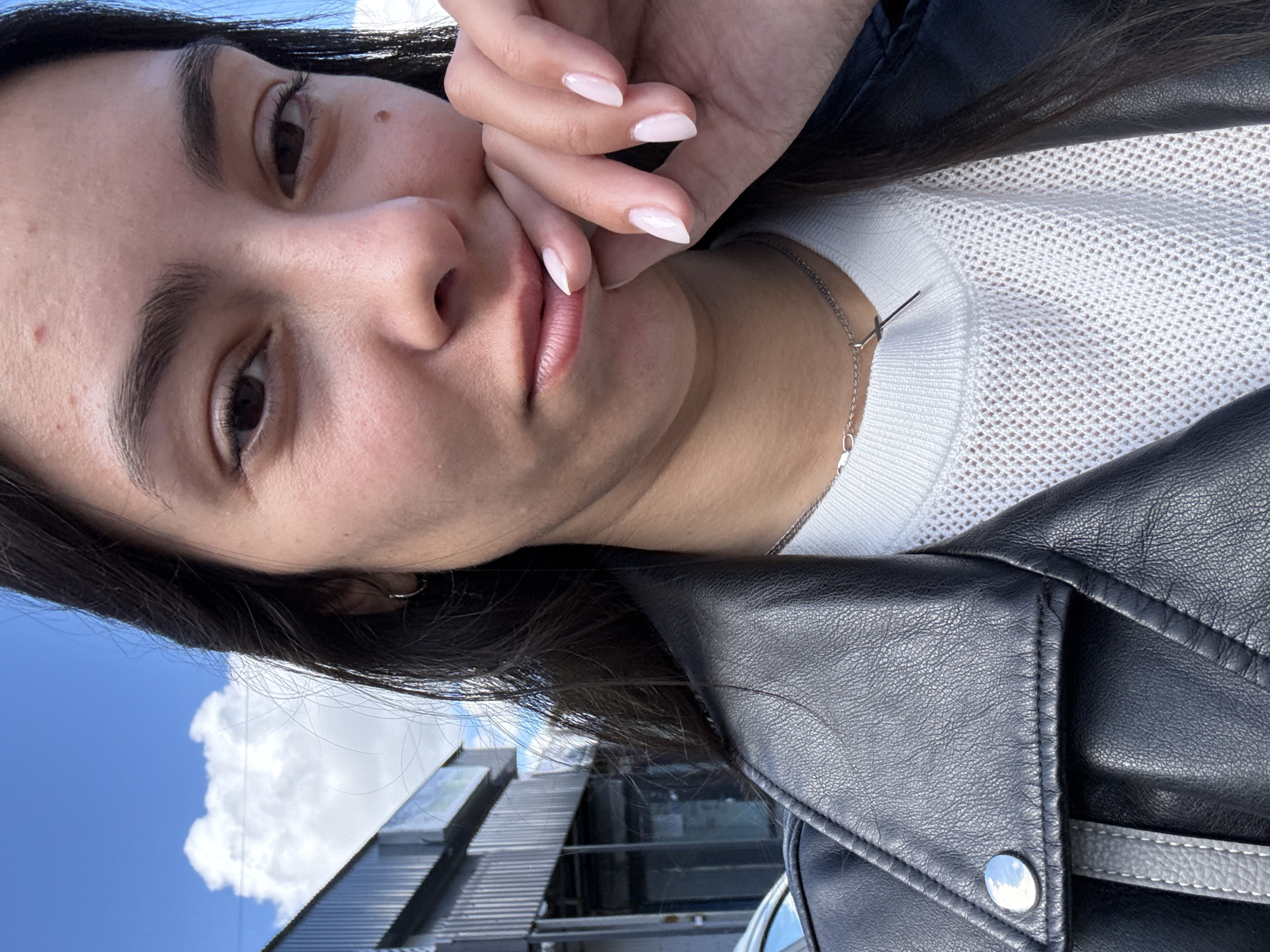 A selfie, angled to the left, features a woman with brown hair, light skin, and brown eyes. She is wearing a white knit sweater and a black leather jacket. Her hand is near her face. She is wearing a silver necklace with a cross. The background includes a building, blue sky, and white clouds. Based on the location information, this is likely in Chaikovsky, Russia.