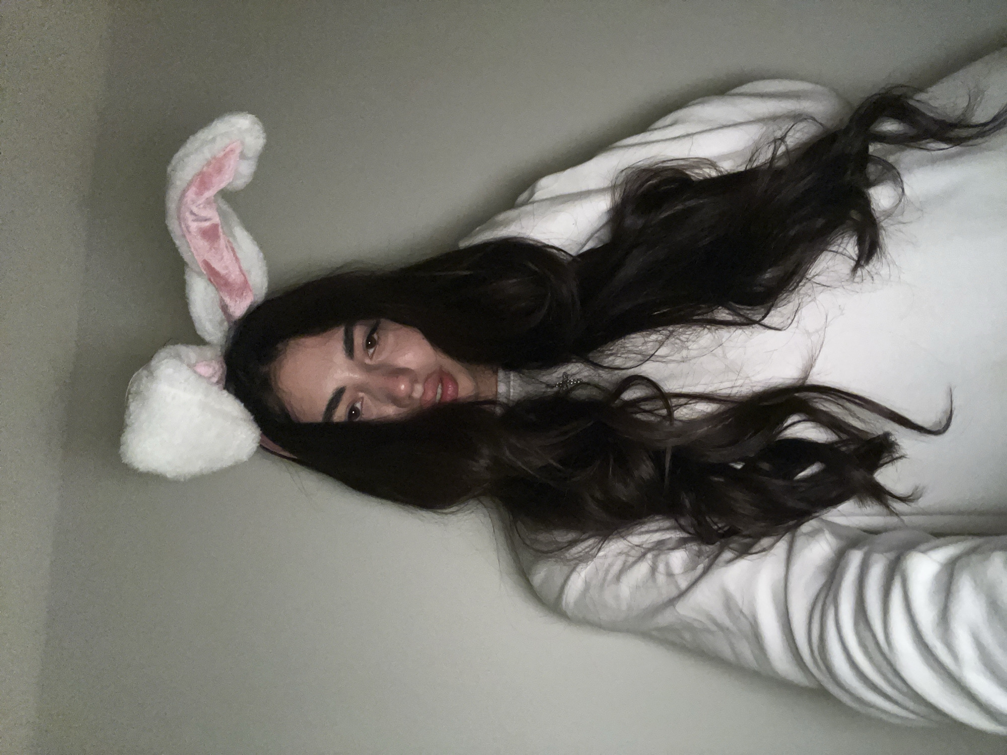 A young woman with long, dark, wavy hair and bunny ears is posing for a selfie against a plain, light-colored wall. She is wearing a white sweatshirt. Her facial expression is somewhat somber, and she is touching her hair on one side. The lighting in the image is dim and casts shadows. There is no indication of the location being Chotěšov, Czechia.