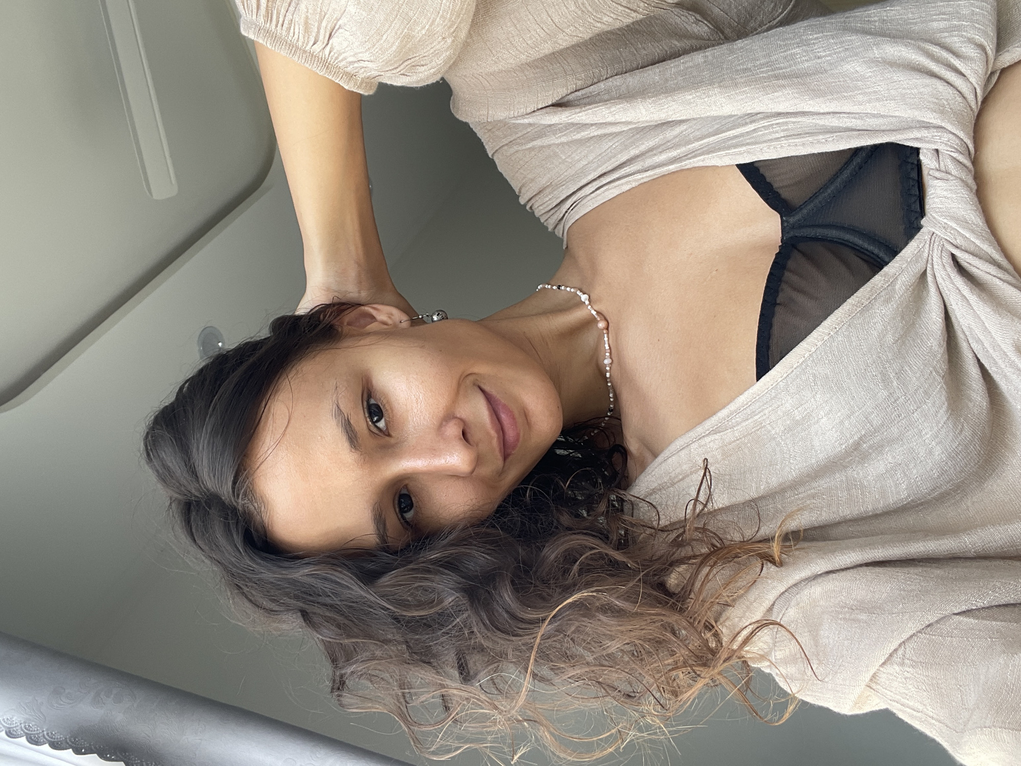 A low-angle shot captures a young woman from the chest up, with the image rotated 90 degrees to the left. She appears to be lying down or reclining, looking directly at the camera with a gentle, inviting smile. Her long, dark, curly hair with lighter brown highlights frames her face and cascades around her head. She has dark eyes and well-defined eyebrows.
She is wearing a light-colored, possibly beige or cream, textured garment that drapes loosely over her upper body, revealing a black sheer-mesh bra underneath. A delicate beaded necklace, featuring small white and pinkish beads, adorns her neck, and a silver earring is visible in her left ear. Her skin tone is warm, appearing light to medium brown.
The background is simple and out of focus, consisting of light-colored, possibly white or light grey, surfaces, suggesting an indoor or shaded environment. There are indistinct lines and shapes in the background, including what might be a bed headboard or a smooth wall. The photo captures a moment of casual repose in Mũi Né, Vietnam. No visible text is present.