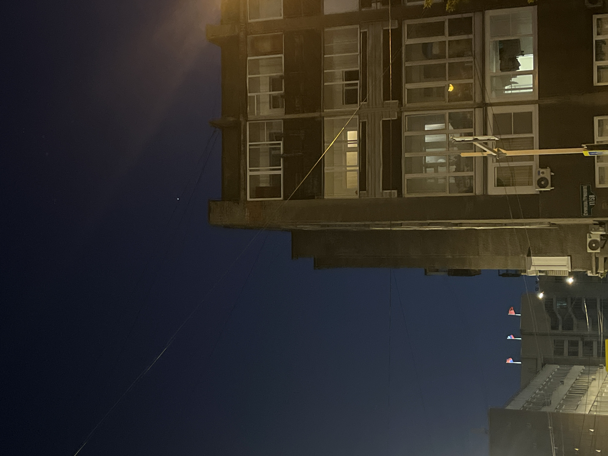 The image shows an upside-down view of a cityscape at night. A multi-story building with numerous windows dominates the upper-right portion of the frame. A street sign is visible, reading "СУРИКОВА/Тургенева 17/58". This indicates a location at the intersection of Surikova and Turgeneva streets, and building number 17/58, which is consistent with a location in Krasnoyarsk, Russia. Power lines run across the dark blue sky on the left side of the image. Further in the distance, another building is partially visible. The overall scene is urban and depicts a nighttime environment.