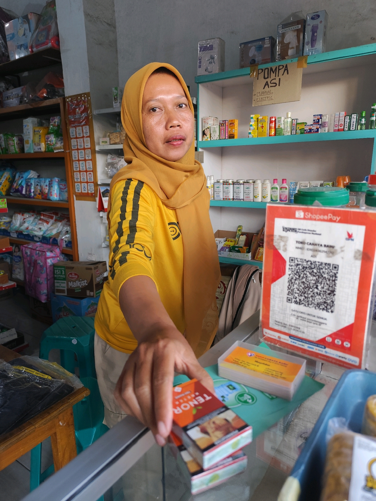 A woman wearing a yellow hijab and a yellow t-shirt with black stripes on the sleeves is standing behind a counter in a small shop. She is holding a pack of cigarettes and reaching out towards the camera. On the counter in front of her is a ShopeePay QR code display, a stack of business cards, and a blue tray containing various items. Behind her, shelves are stocked with various products, including baby items, boxes, and bottles of what appear to be toiletries or medicines. The shop appears to be in Bambang Lipuro, Indonesia, as indicated by a sign on the wall. The lighting is bright, suggesting it is daytime.