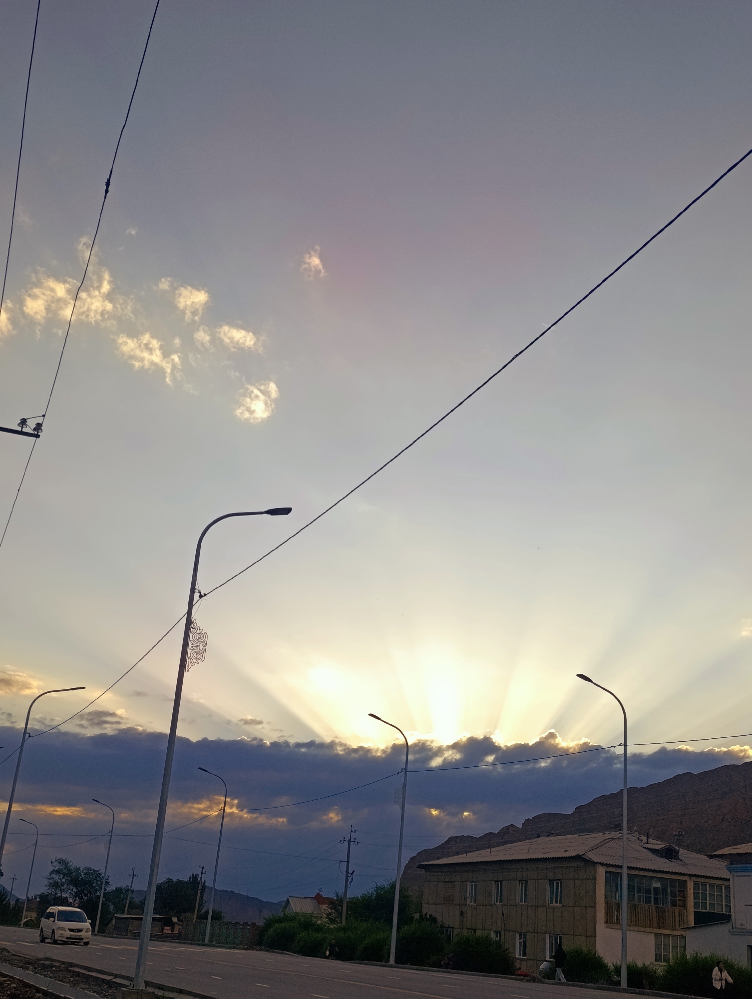 The image depicts a street scene in Naryn City, Kyrgyzstan, during sunset. The sky is illuminated by crepuscular rays emanating from behind dark clouds, creating a dramatic effect. The sun is low on the horizon, casting a warm glow. Several streetlights line the road, some with decorative elements. A multi-lane asphalt road occupies the foreground, with a white van driving on it. To the right of the road, a two-story building with a light-colored facade and a dark roof is visible, partially obscured by trees and bushes. Mountains form the backdrop, suggesting the city's mountainous terrain. A few individuals are present on the sidewalk and near the building. Power lines are strung across the sky, adding to the urban infrastructure. The overall atmosphere is serene, characteristic of a city winding down for the evening.