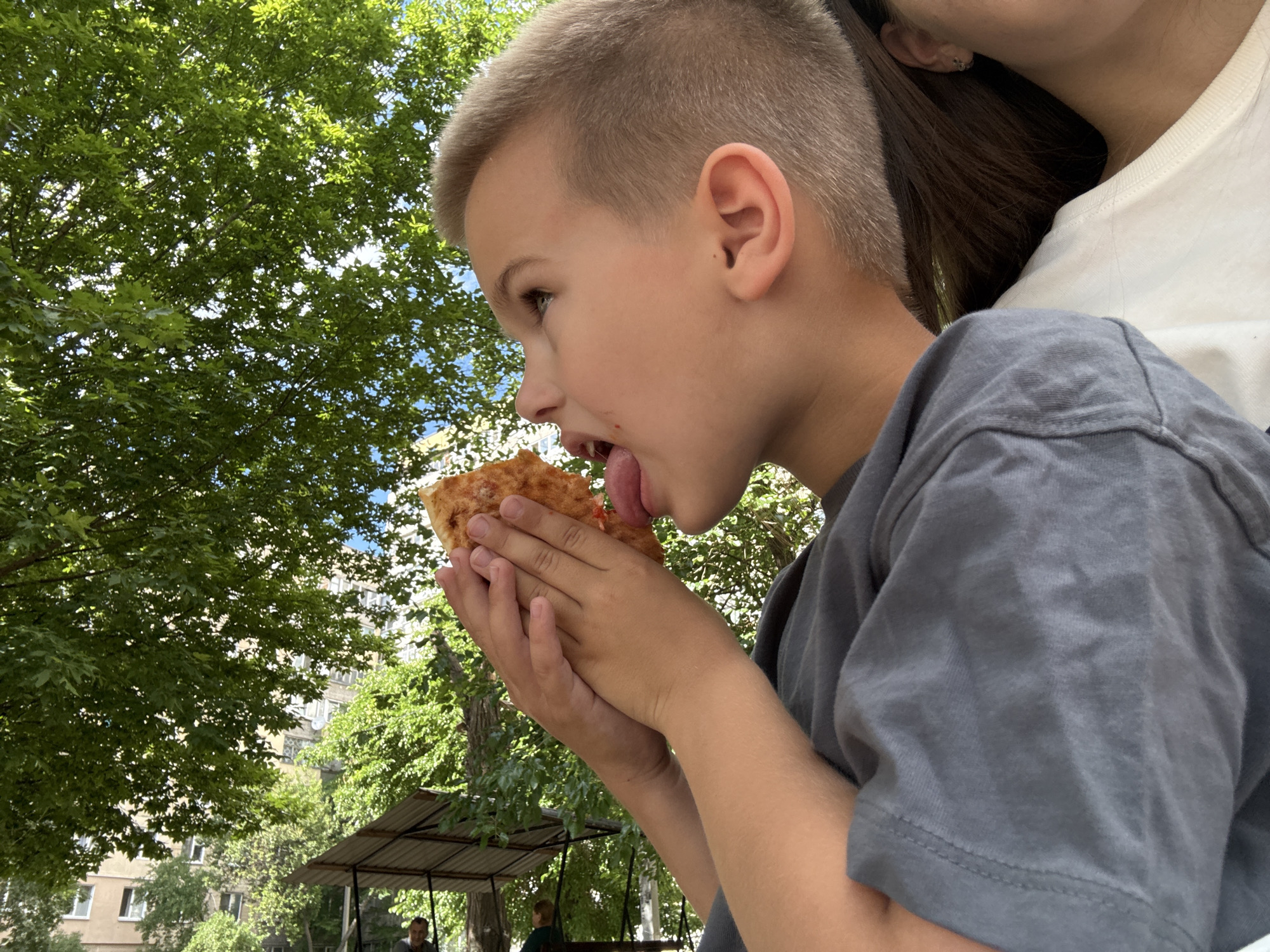 A young boy is eating a slice of pizza outdoors. He is wearing a gray t-shirt and his hair is short and blonde. His tongue is sticking out as he takes a bite of the pizza. He is holding the pizza with both hands. In the background, there are green trees and buildings, suggesting an urban environment. The scene appears to be in a park or courtyard, with a small covered structure visible. Based on visual cues like the architecture of the buildings and the presence of mature trees, this location is consistent with the city of Dnipro, Ukraine.