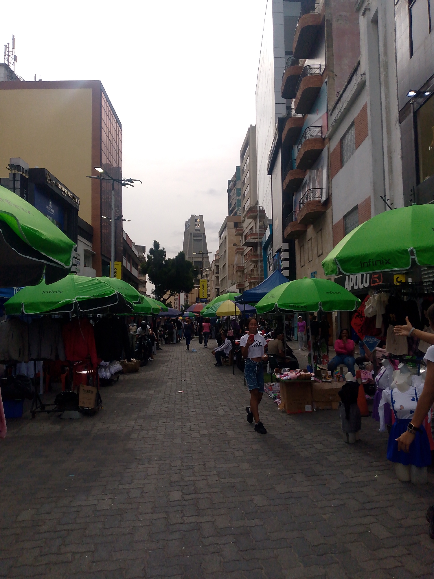The image shows a street scene in Caracas, Venezuela, likely a pedestrianized shopping area. The street is paved with interlocking bricks. There are vendors on both sides of the street, selling goods from under green umbrellas with brand names (Infinix, Apolu 51). A woman in a white t-shirt and denim shorts walks toward the camera while holding a phone. Other pedestrians walk further down the street. Buildings line the street, with one in the distance being a tall, distinctively shaped structure with a cross at the top. Some buildings are multi-story with balconies. Street lights are also visible. Clothes, accessories and other goods are on display.