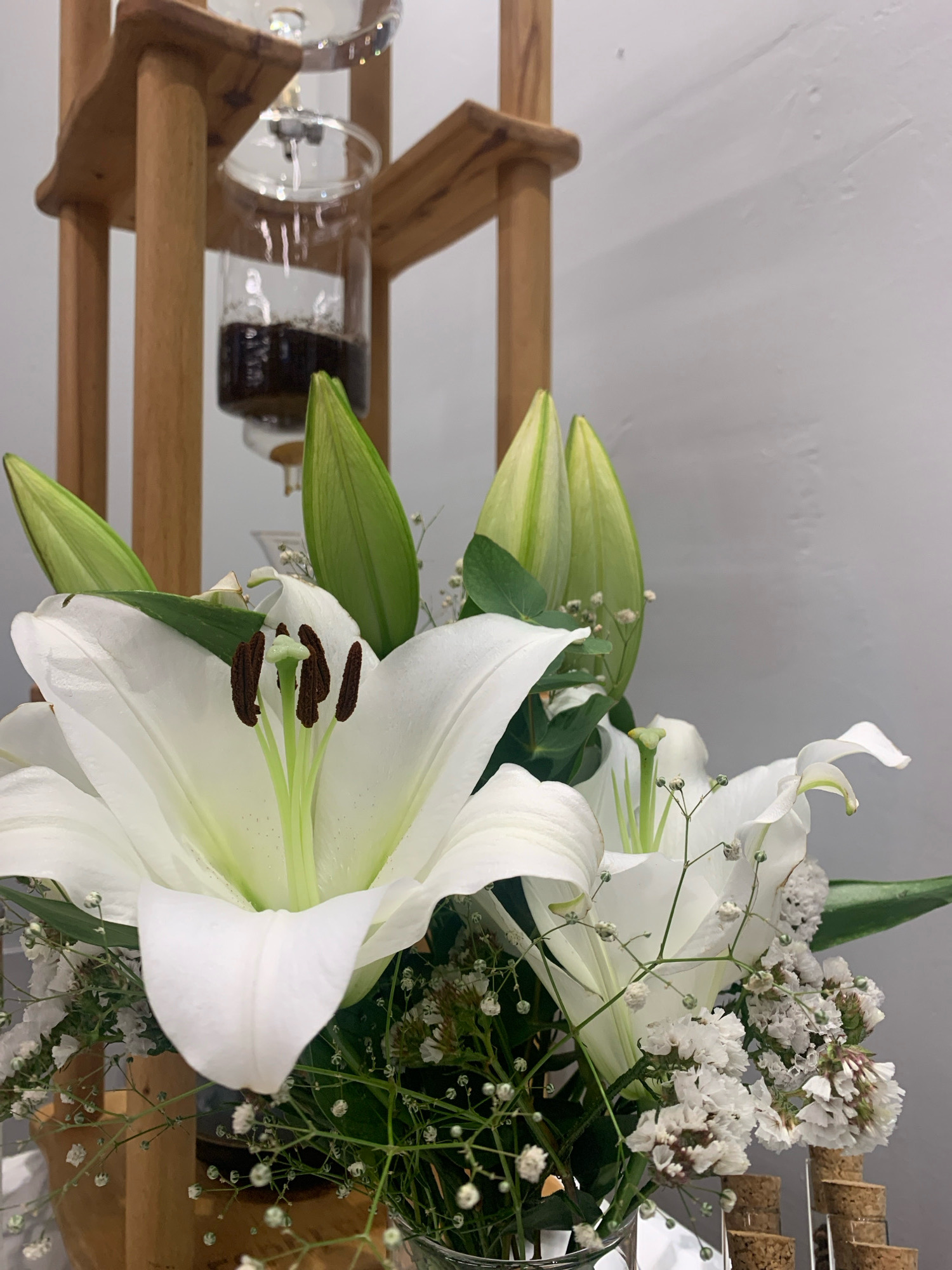 The image shows a close-up of a white lily in bloom, with several unopened buds behind it. The flower has prominent brown anthers and a pale green stigma. It is part of a bouquet that also includes small white flowers and greenery. In the background, there is a wooden structure with glass vessels containing a dark liquid, possibly a coffee maker. The setting appears to be indoors, with a plain gray wall in the background. The overall impression is one of a still life arrangement. There are no people or animals visible in the image, and no specific indication of the city of Beyoğlu, Turkey.