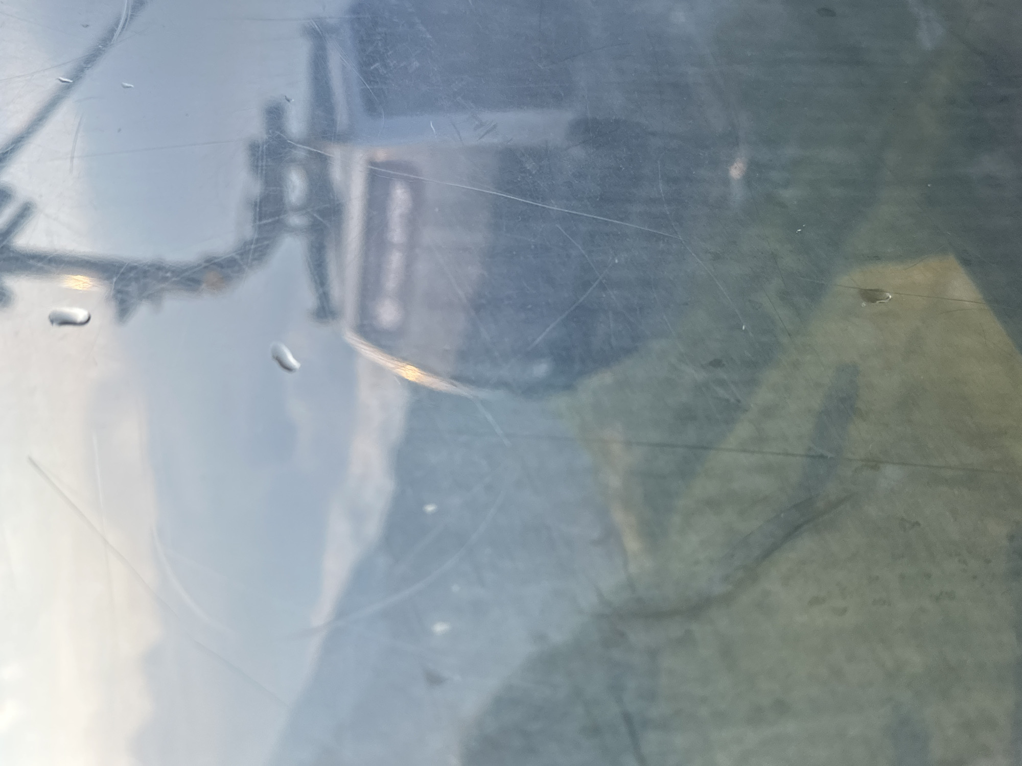 The image shows a blurry view through a window, likely from a cable car, overlooking a mountainous landscape. A cable car cabin and its supporting structure are visible in the upper right portion of the frame, slightly out of focus. The lower left of the image reveals a green, mountainous terrain, also blurred by the window. Water droplets and scratches are present on the glass, further obscuring the view. The sky appears overcast. The scene is consistent with being in Estosadok, Russia, a popular mountain resort area known for its cable car systems and scenic views.