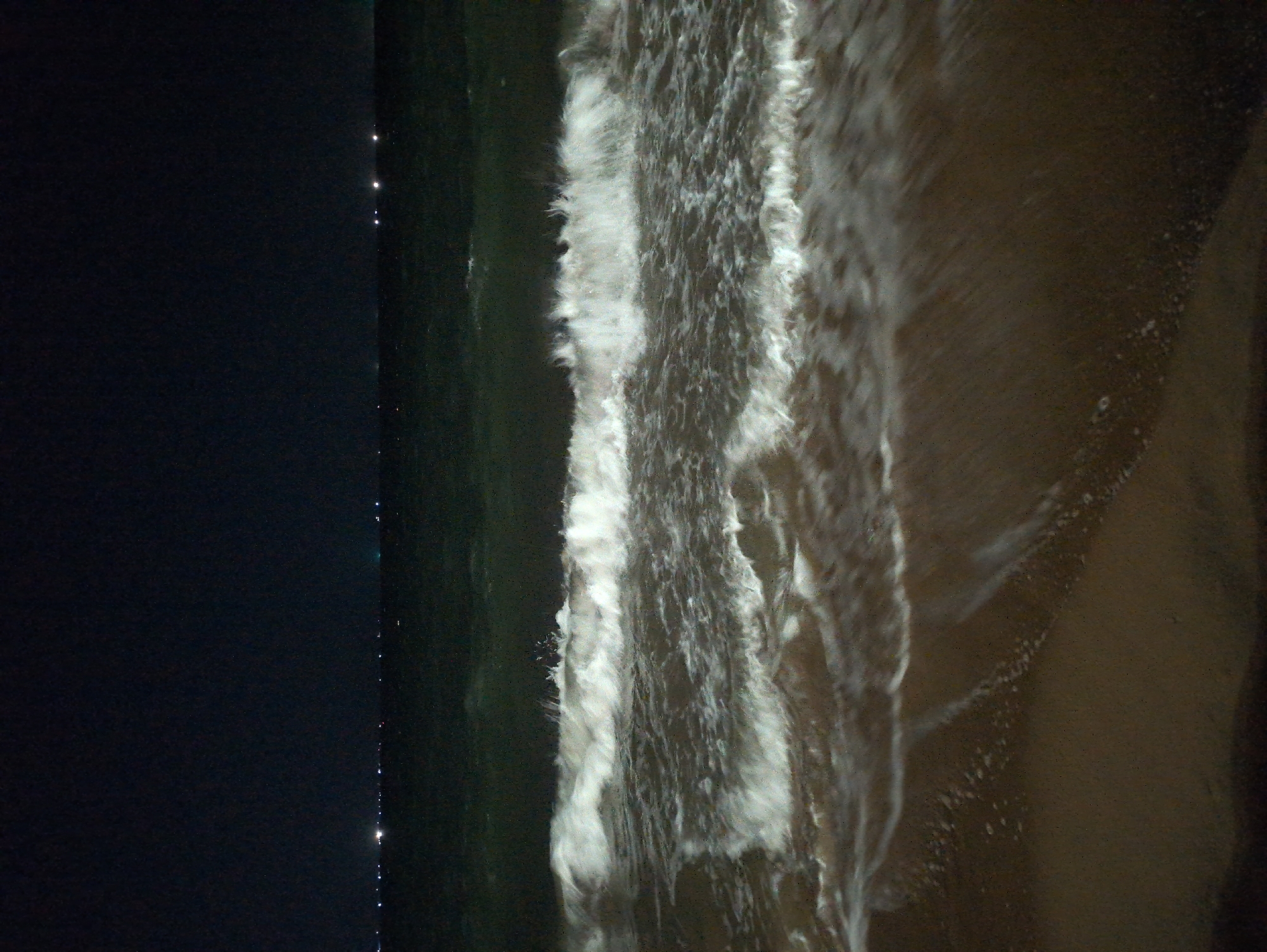 The image shows an aerial view of a beach at night, possibly in South Quy Nhon Ward, Vietnam. A dark body of water borders the left side of the frame, with faint light reflections on its surface. White, foamy waves surge toward the right, where the shoreline meets a dark sand beach. The scene is dimly lit.