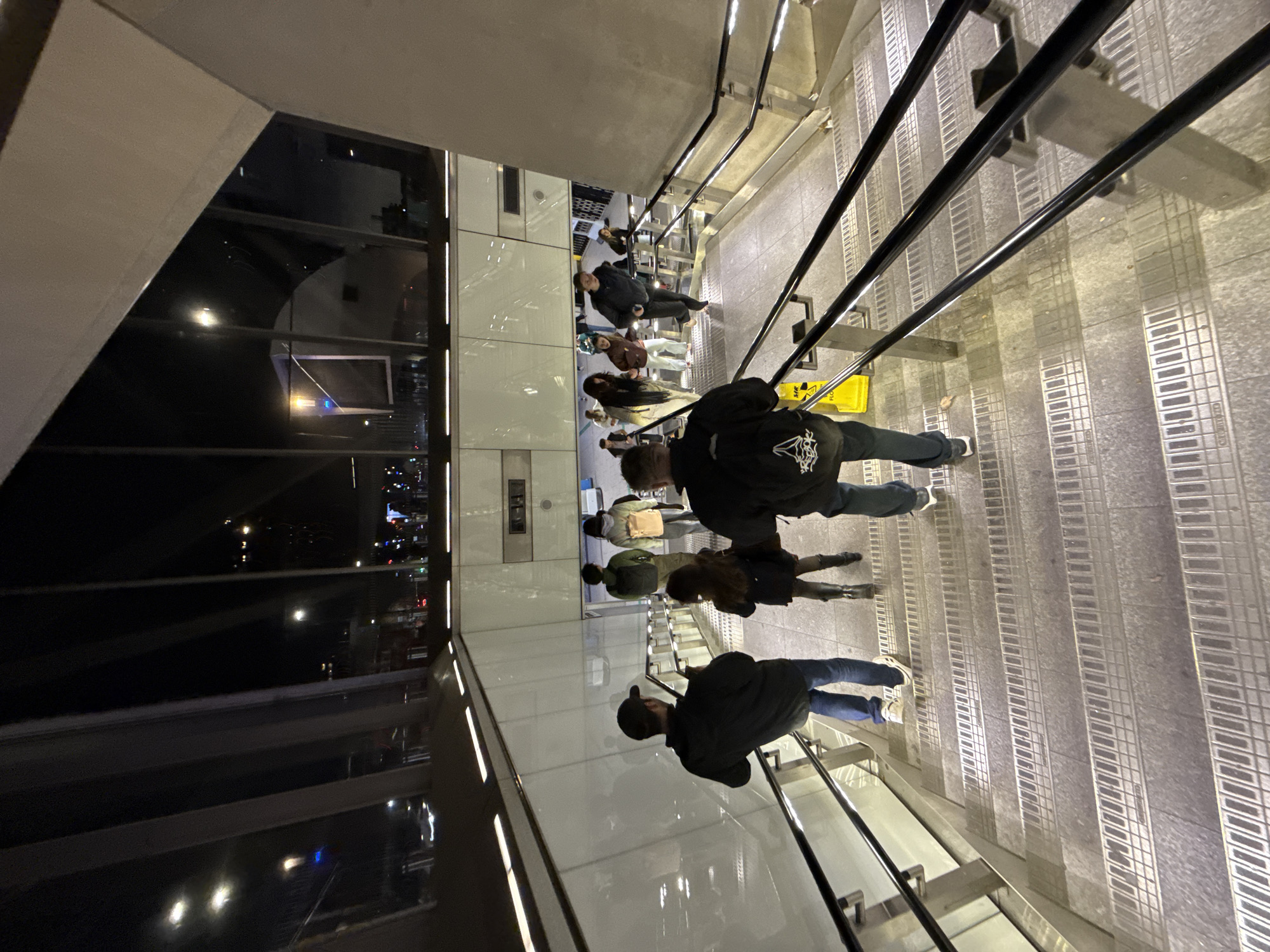 The image shows a downward view from an elevated position within a modern, brightly lit indoor space, likely a public building in London, United Kingdom. The scene features a stairway with metal handrails, descending towards what appears to be a lower level. Several people are walking down the stairs in a staggered fashion. The environment includes large glass windows that offer a view of the cityscape at night, illuminated by artificial lights. The architectural design is sleek and contemporary, and the scene suggests a bustling, urban environment.