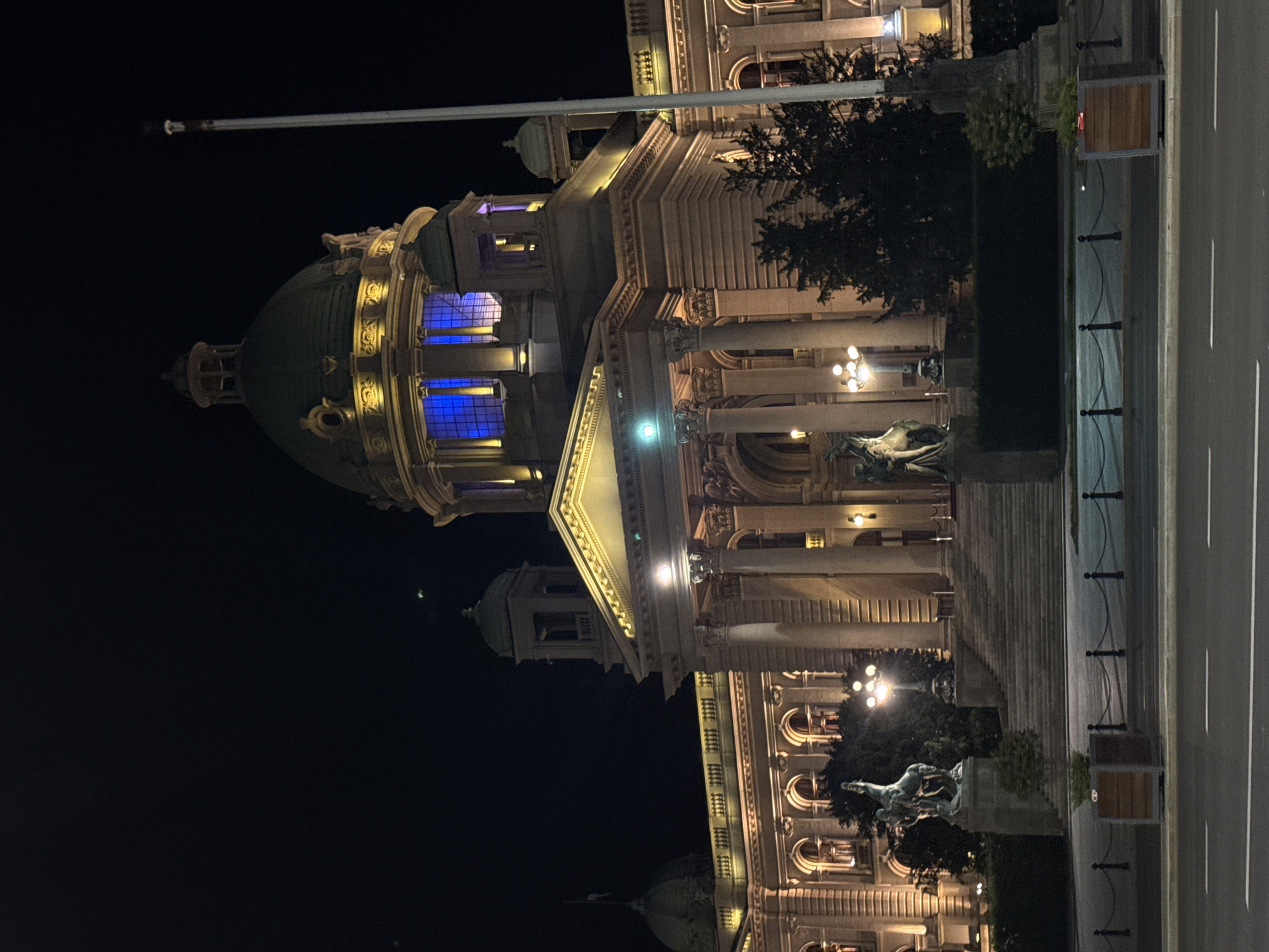 The image is a night view of the National Assembly Building in Belgrade, Serbia, also known as the Dom Narodne Skupštine. The building, illuminated with warm light, features a central dome with blue lighting emanating from within. Classical architectural elements, including columns and a triangular pediment, are visible. A flag pole extends from the building's roofline. In front of the assembly building is a road and a fence with ornamental posts. Below, statues are present. The night sky is dark, contrasting sharply with the illuminated building. The image appears to be rotated 90 degrees counterclockwise.