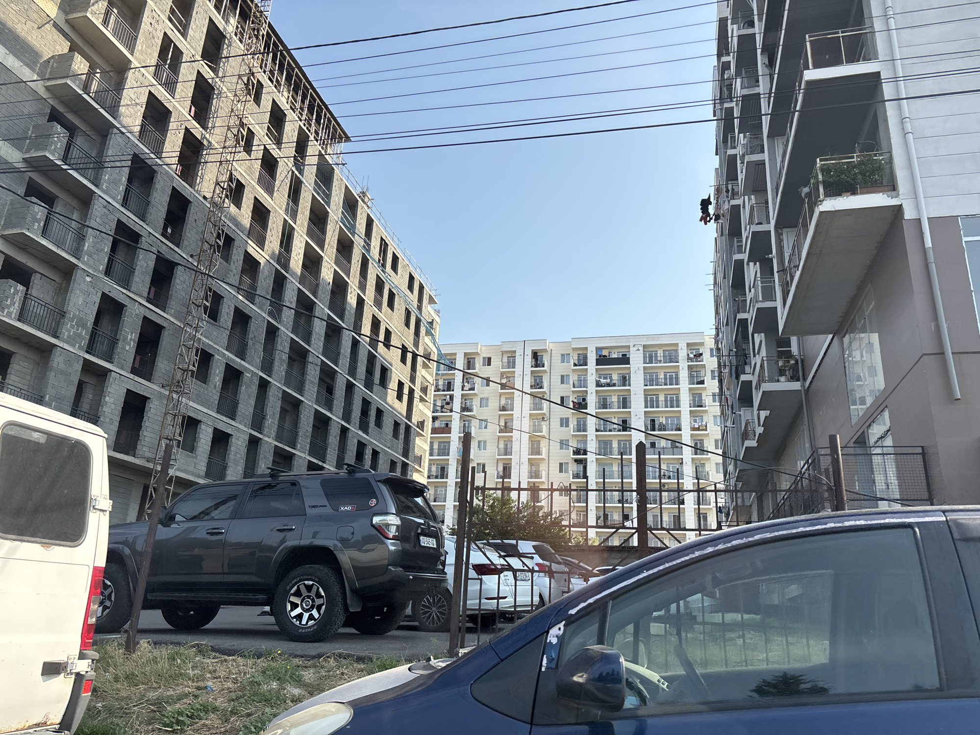 The image shows a street scene in Tbilisi, Georgia, with a focus on residential buildings under construction and completed apartment blocks. In the foreground, a dark blue car is partially visible, with its side and windshield in view. To the left, a white van is parked. Behind these vehicles, a dark gray SUV is parked, displaying a "TRD PRO" decal on its rear panel.
The background is dominated by several multi-story buildings. On the left, a tall building under construction is visible, made of concrete blocks with exposed rebar and metal scaffolding. In the center, a lighter-colored apartment building with numerous balconies and windows is seen. To the right, another building, also with balconies, stands.
Power lines are strung across the upper portion of the image, contributing to the urban atmosphere. One detail of note is laundry hanging from a balcony on the right-hand building, with two items of clothing suspended by clothespins. A person appears to be climbing on the side of the construction building on the left, using what looks like a ladder or scaffolding. The sky is clear and blue.