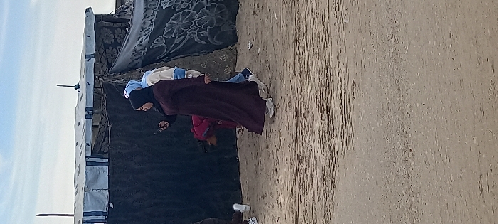 An outdoor scene in Khan Yunis, Palestinian Territory, depicts several individuals on a sandy ground in front of makeshift structures under a light blue sky. In the foreground, a female wearing a dark, long abaya and a black hijab stands in profile, facing left, looking down at a dark-colored smartphone held in her left hand. She wears white sneakers. Partially obscured behind her and to the right, another individual with a light blue hijab and a light-colored top with blue accents is visible, walking away to the right, wearing blue and white sneakers. Further back, positioned between these two figures, a person dressed in a red garment is bent over, facing the ground. To the far left, a white sneaker and lower leg of another individual are partially visible. The background features temporary structures constructed from various materials, including a white tarp with prominent blue stripes forming a roofline, and darker, patterned fabrics or blankets serving as walls. Vertical poles extend upwards from these structures. The ground is predominantly light brown sand or packed dirt with subtle textural variations.