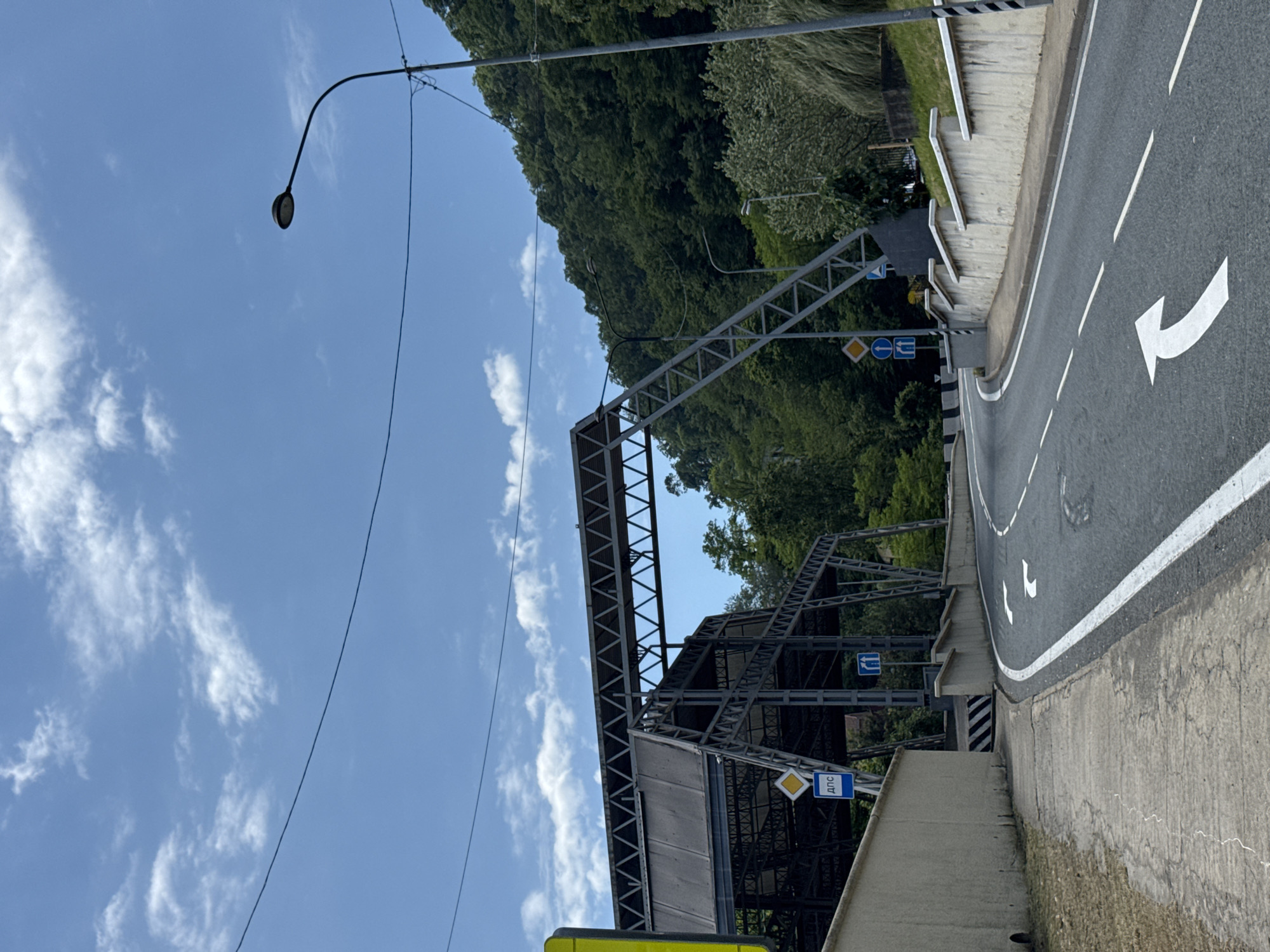 A public road curves to the right, bordered by a concrete wall on the right and a sidewalk on the left. The road is marked with white lane lines and directional arrows. In the background, a large metal structure, possibly an overpass or part of a building, is visible against a backdrop of lush green trees and a blue sky with scattered clouds. A streetlamp stands on the right side of the road. The scene appears to be in Sochi, Russia, based on the presence of the structure and the natural environment.