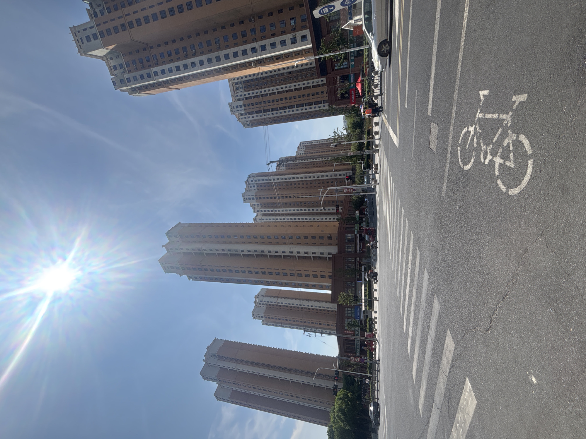 This image captures a street scene in Suzhou Industrial Park, China, under a bright, sunny sky with prominent lens flare. Multiple tall, orange-brick residential buildings with white accents line the background. In the foreground, a wide asphalt road features a marked crosswalk and a painted bicycle lane symbol. Several cars are visible on the road, including a silver sedan in the right lane and a dark sedan in the left lane. A few pedestrians are present near the crosswalk, and a person on a scooter is also in view. Traffic lights and street lamps are positioned at the intersection. The overall environment suggests a modern urban setting with residential and commercial elements.