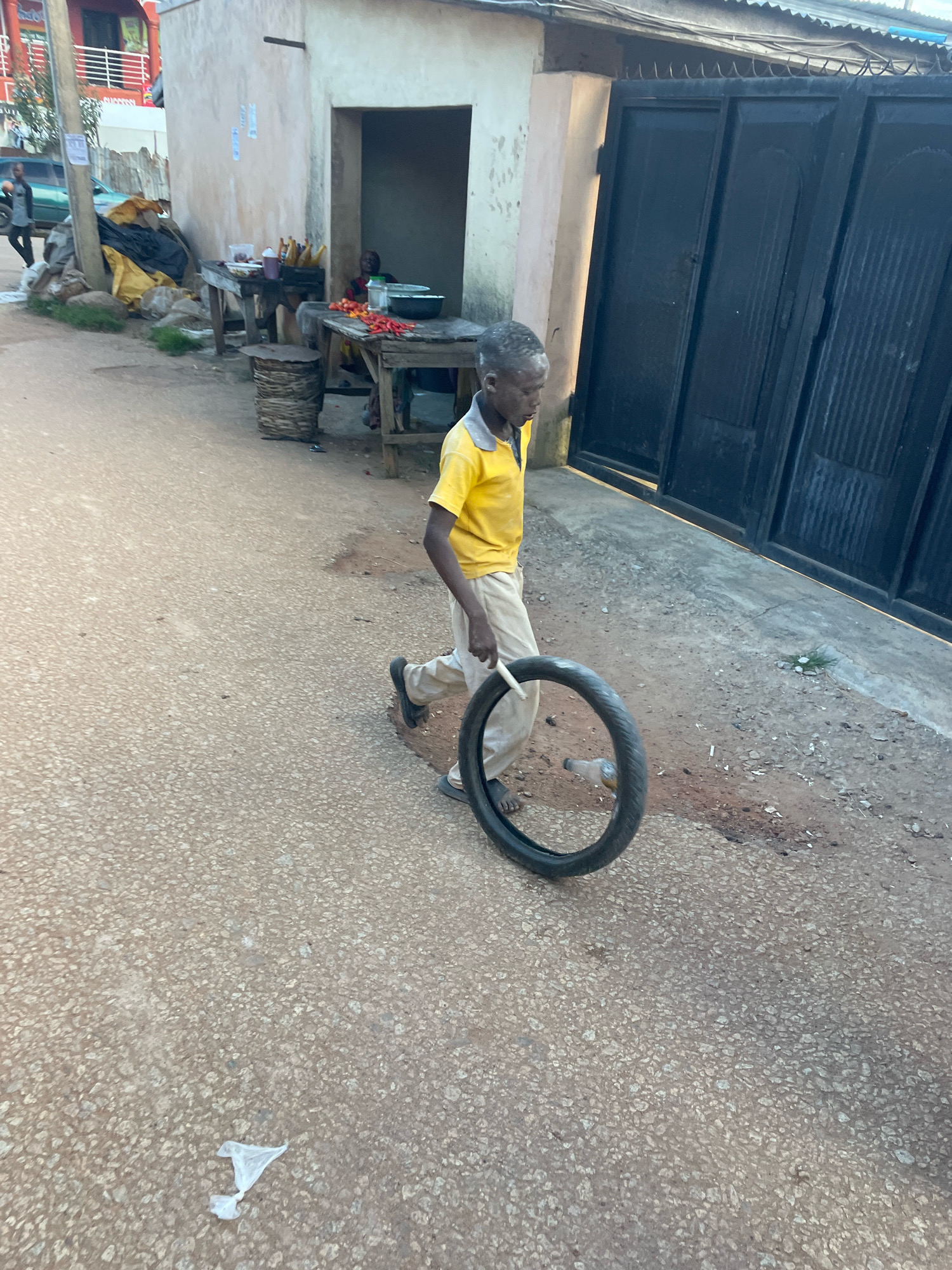 In Jos, Nigeria, a young boy in a yellow shirt and beige pants rolls a tire along a dirt road. He is walking in front of a building with a dark metal gate, which appears to be a shop. To the left of the building, a street vendor's stall is visible, with tables laden with produce like peppers and corn. The road surface is a mix of gravel and dirt. The scene suggests a common street view in a bustling urban environment.